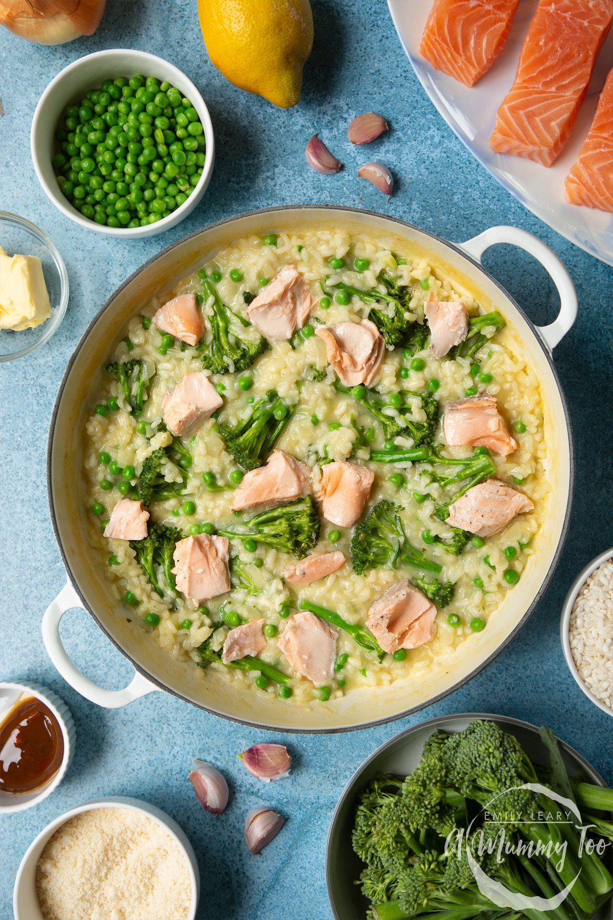 Freshly cooked risotto with peas and Tenderstem broccoli and pieces of salmon flaked on top in a large cream-colour pan. Ingredients to make salmon risotto surround the pan.