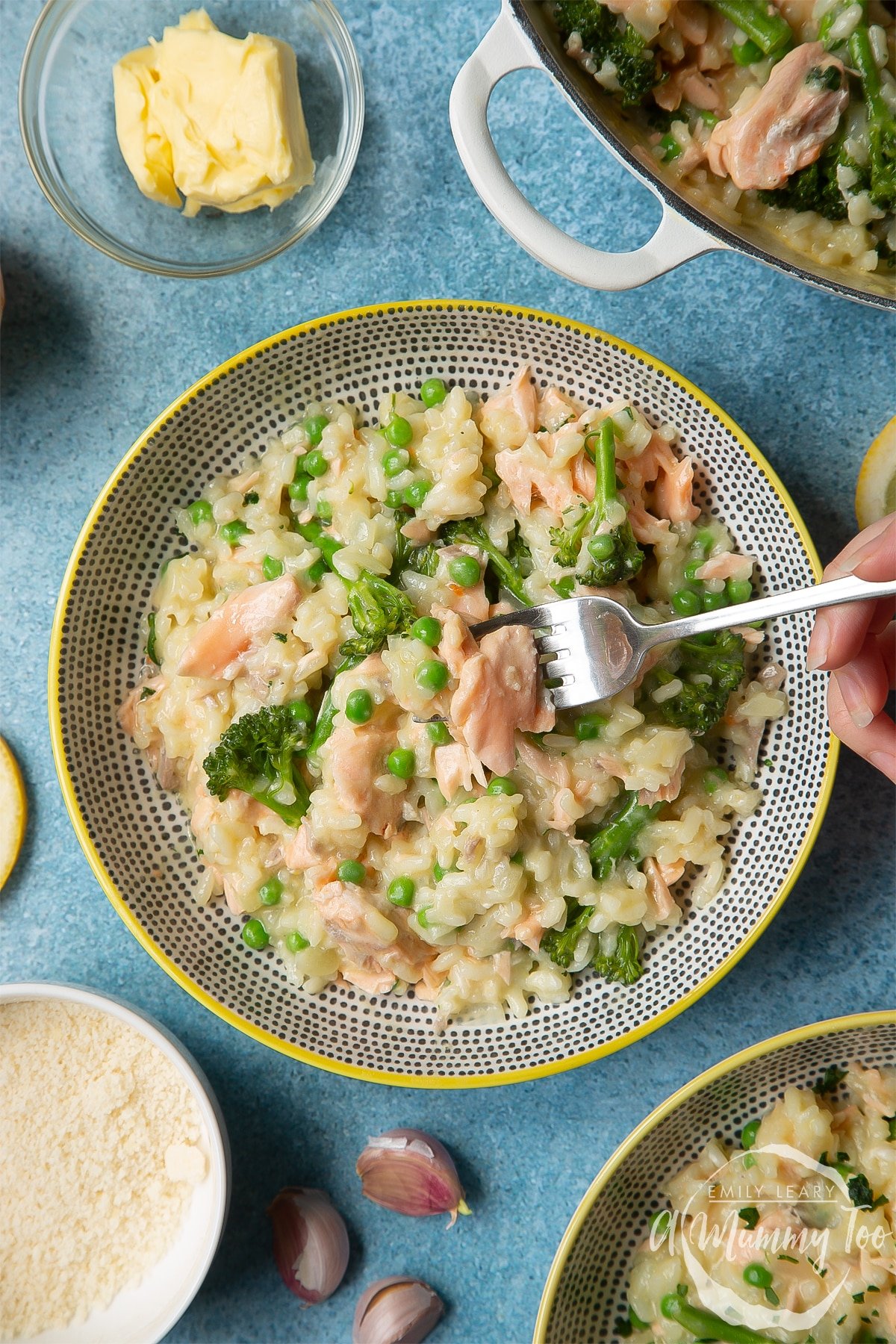 Salmon risotto in a spotted bowl with a yellow rim. A hand holding a fork takes a piece of salmon.