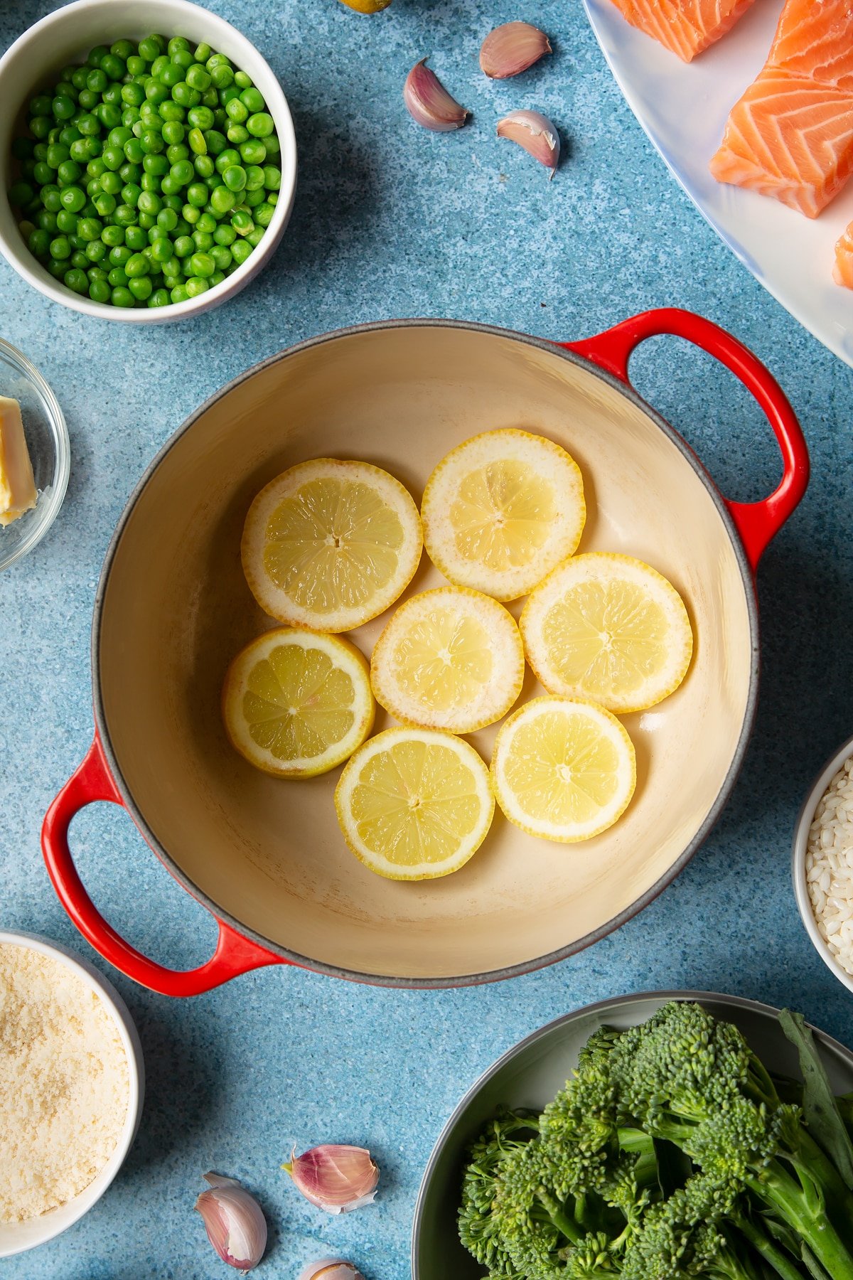 Sliced lemons in the bottom of a red saucepan. Ingredients to make salmon risotto surround the pan.