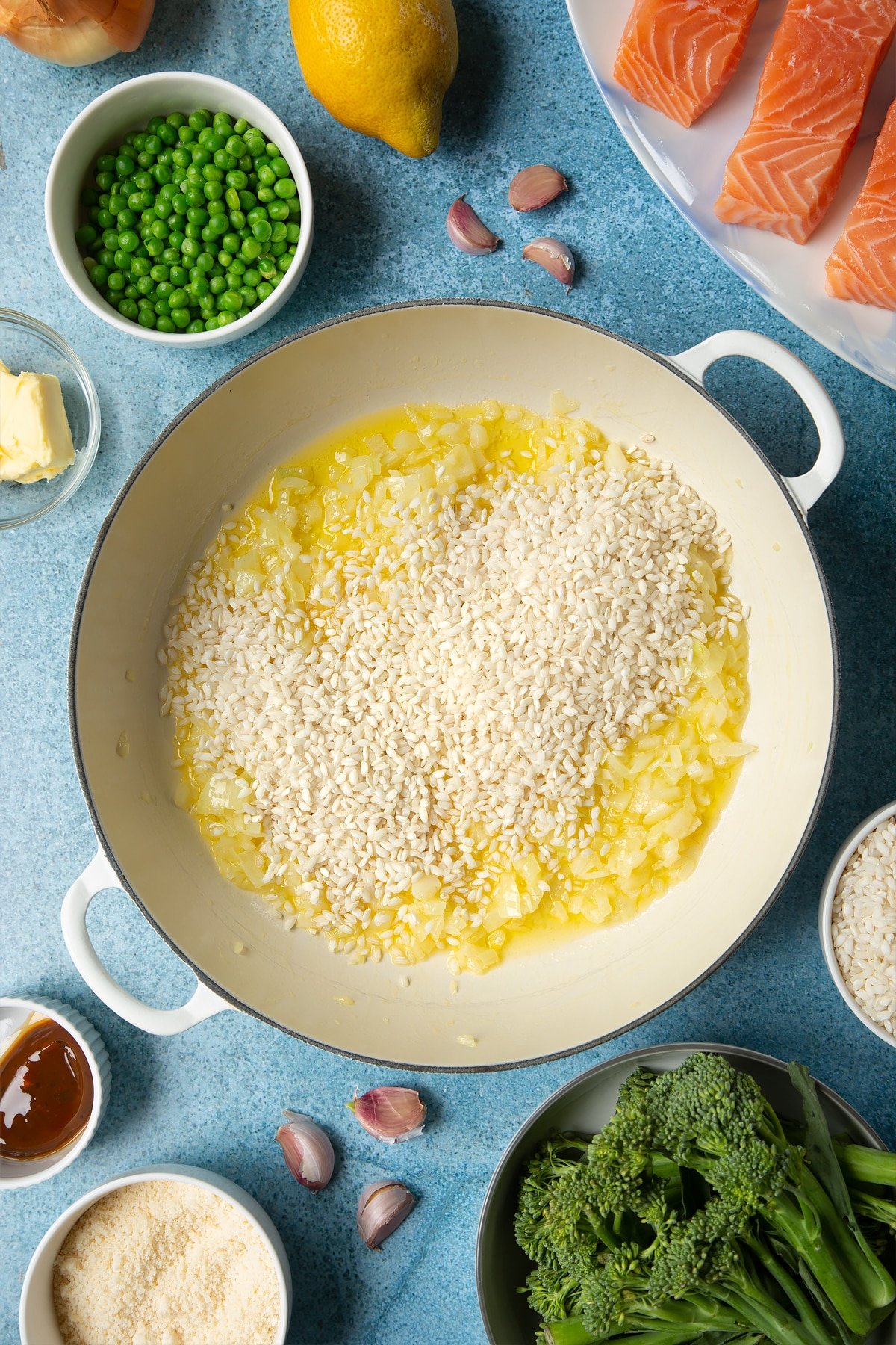 Onions, garlic and butter with arborio rice on top in a large cream-colour pan. Ingredients to make salmon risotto surround the pan.