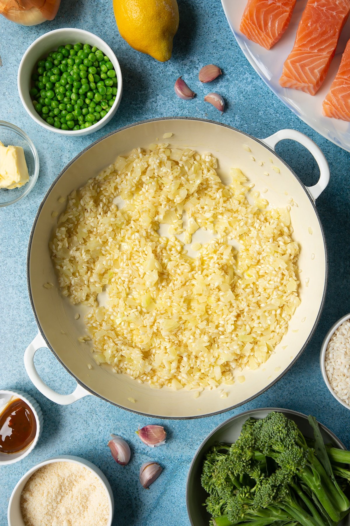 Onions, garlic, butter and arborio rice mixed together in a large cream-colour pan. Ingredients to make salmon risotto surround the pan.