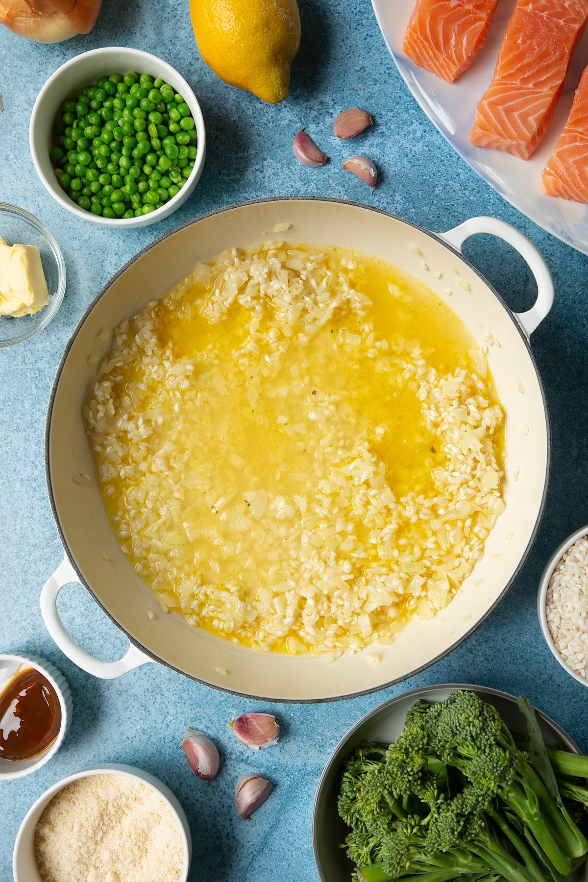 Onions, garlic, butter and arborio rice with stock on top in a large cream-colour pan. Ingredients to make salmon risotto surround the pan.
