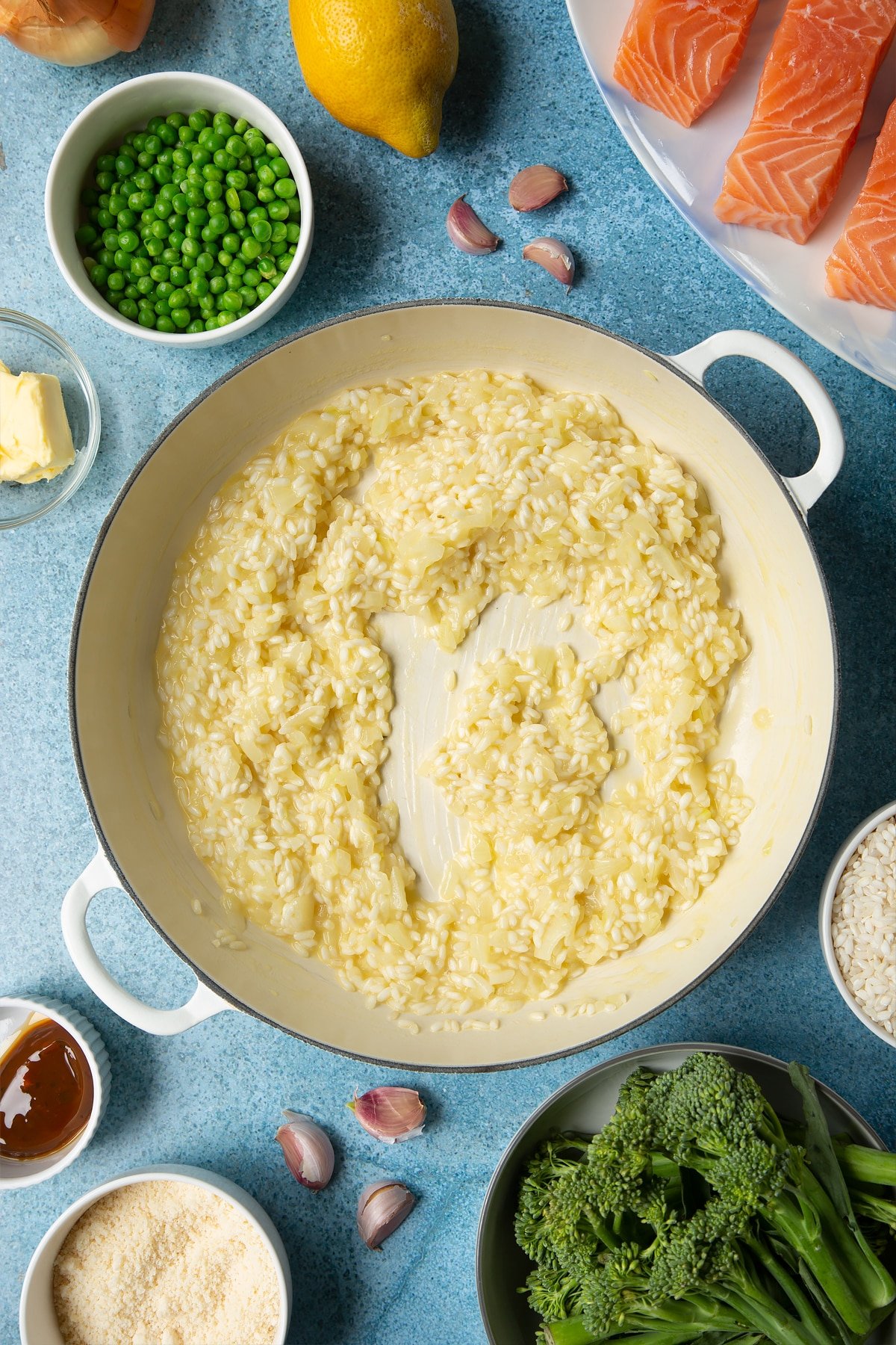 Onions, garlic, butter and arborio rice with stock forming a semi-cooked risotto in a large cream-colour pan. Ingredients to make salmon risotto surround the pan.