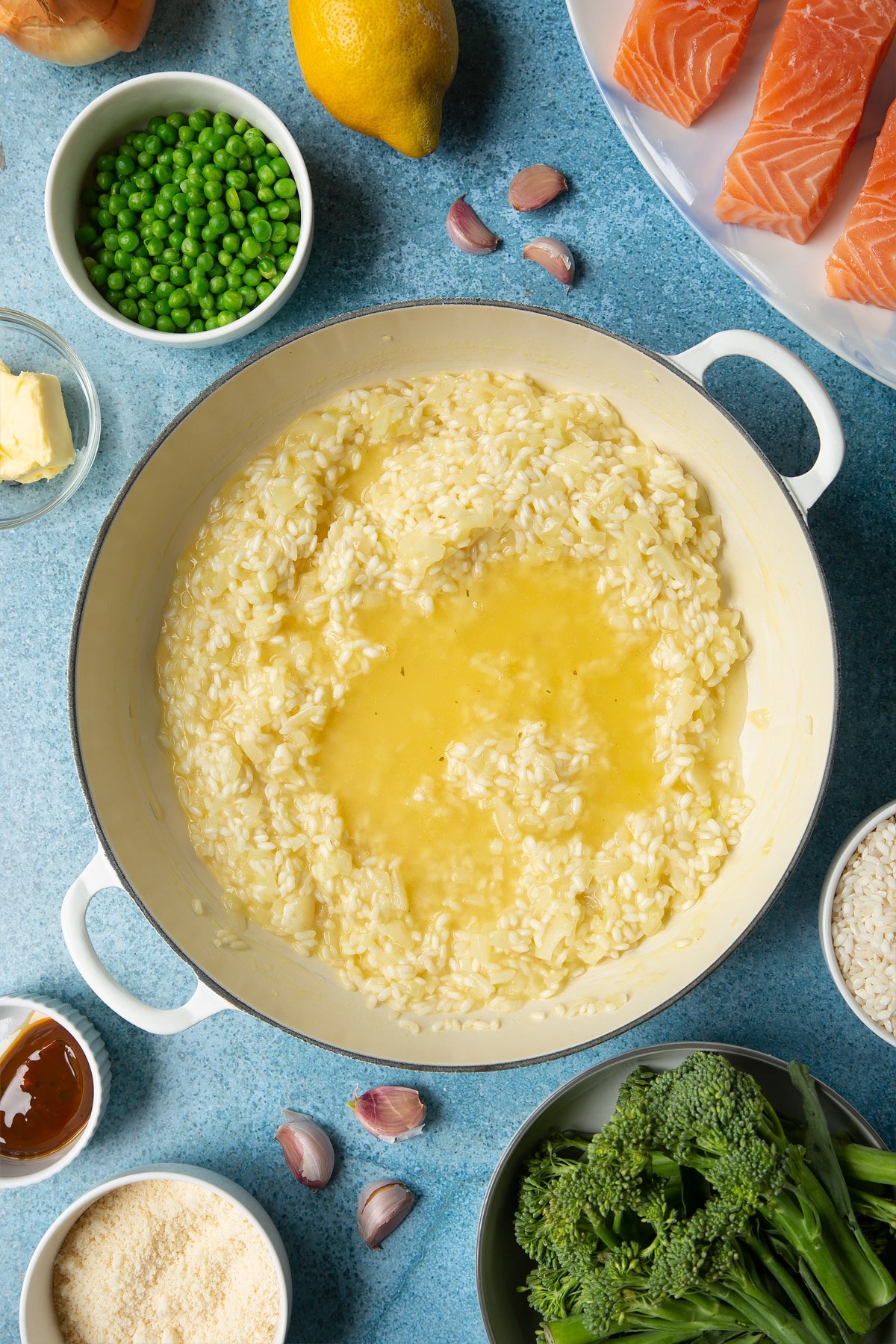 Onions, garlic, butter and arborio rice forming a semi-cooked risotto, with stock on top in a large cream-colour pan. Ingredients to make salmon risotto surround the pan.