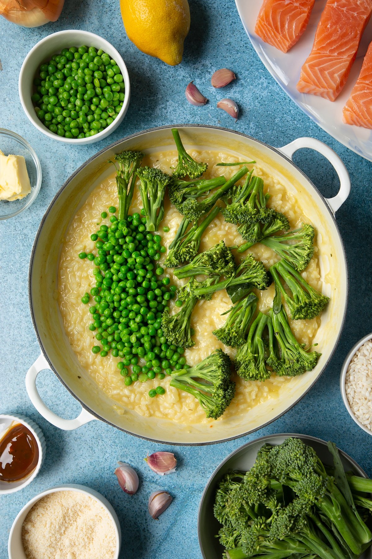 Freshly cooked risotto with peas and Tenderstem broccoli on top in a large cream-colour pan. Ingredients to make salmon risotto surround the pan.