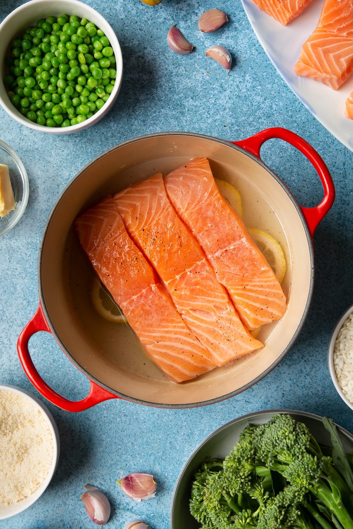 Sliced lemons and hot water in the bottom of a red saucepan with seasoned salmon fillets on top. Ingredients to make salmon risotto surround the pan.