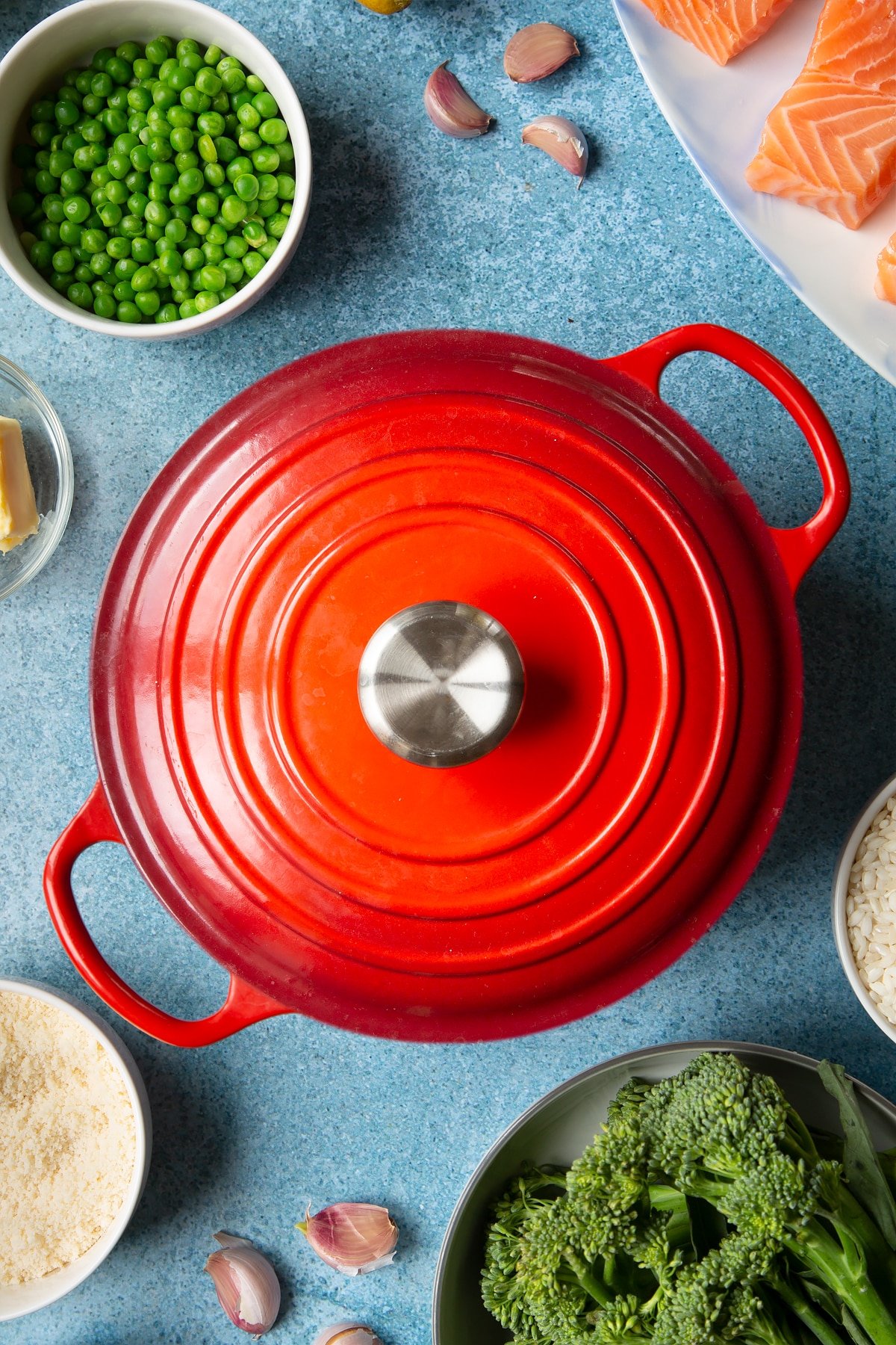 Lidded red saucepan. Ingredients to make salmon risotto surround the pan.