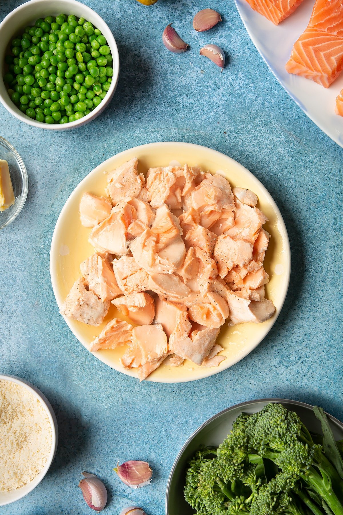 Cooked, flaked salmon on a pale yellow plate. Ingredients to make salmon risotto surround the plate.