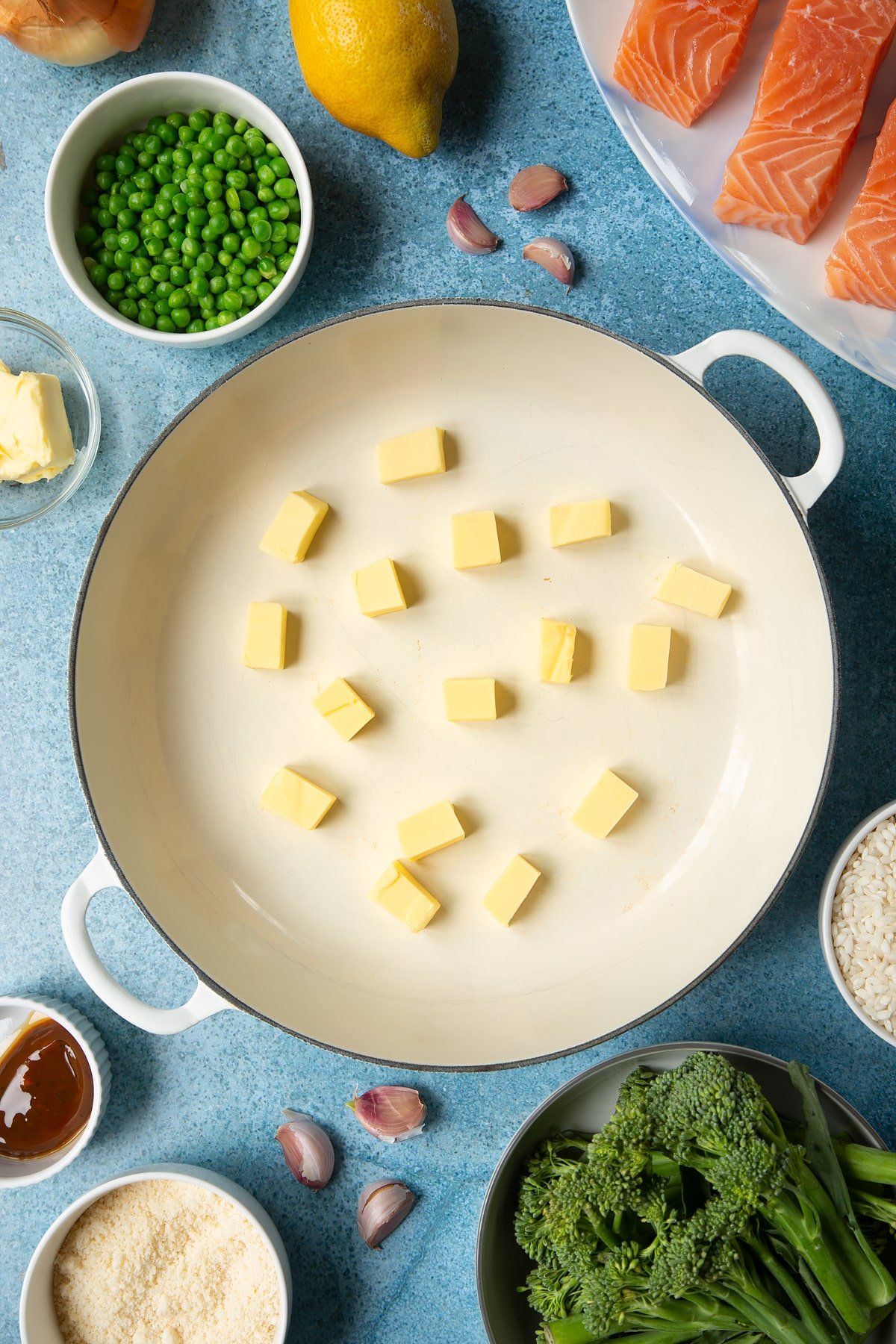 Cubes of butter in a large cream-colour pan. Ingredients to make salmon risotto surround the pan.