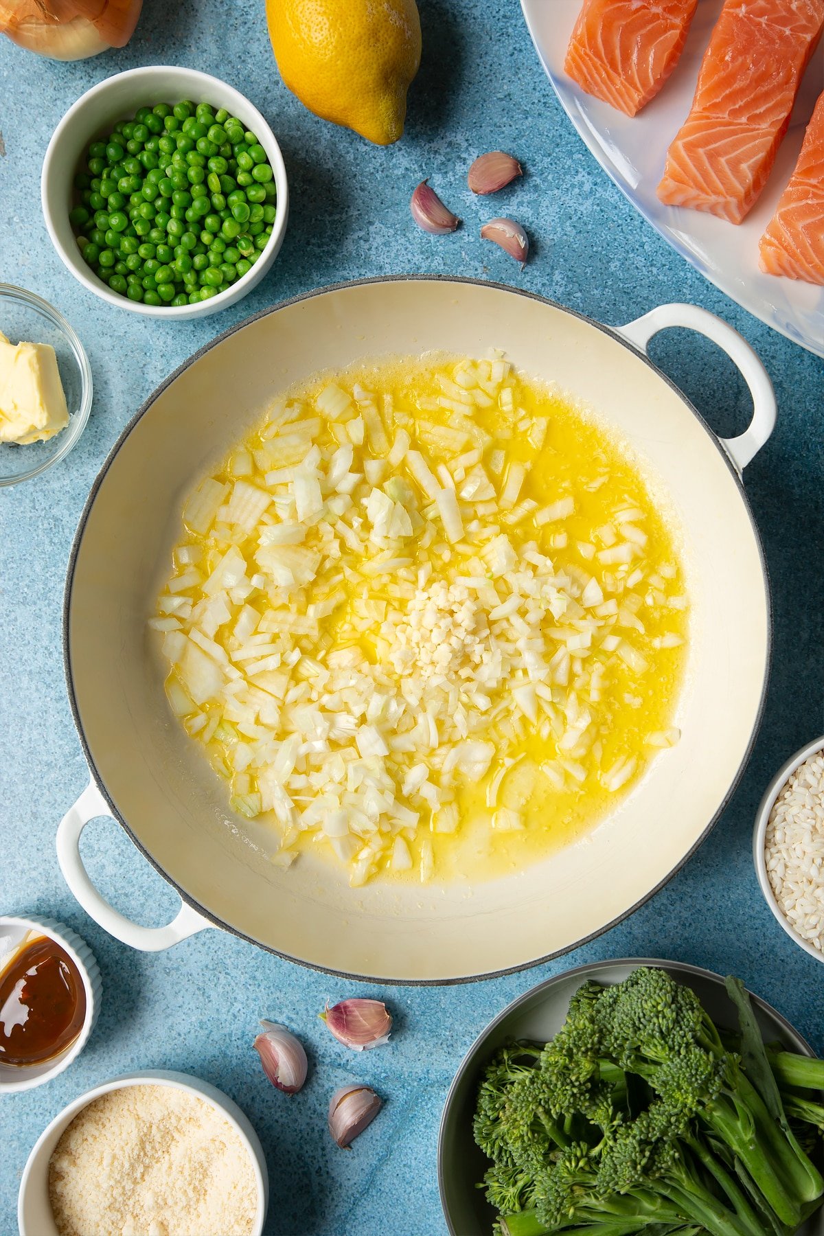 Melted butter, chopped onions and chopped garlic in a large cream-colour pan. Ingredients to make salmon risotto surround the pan.