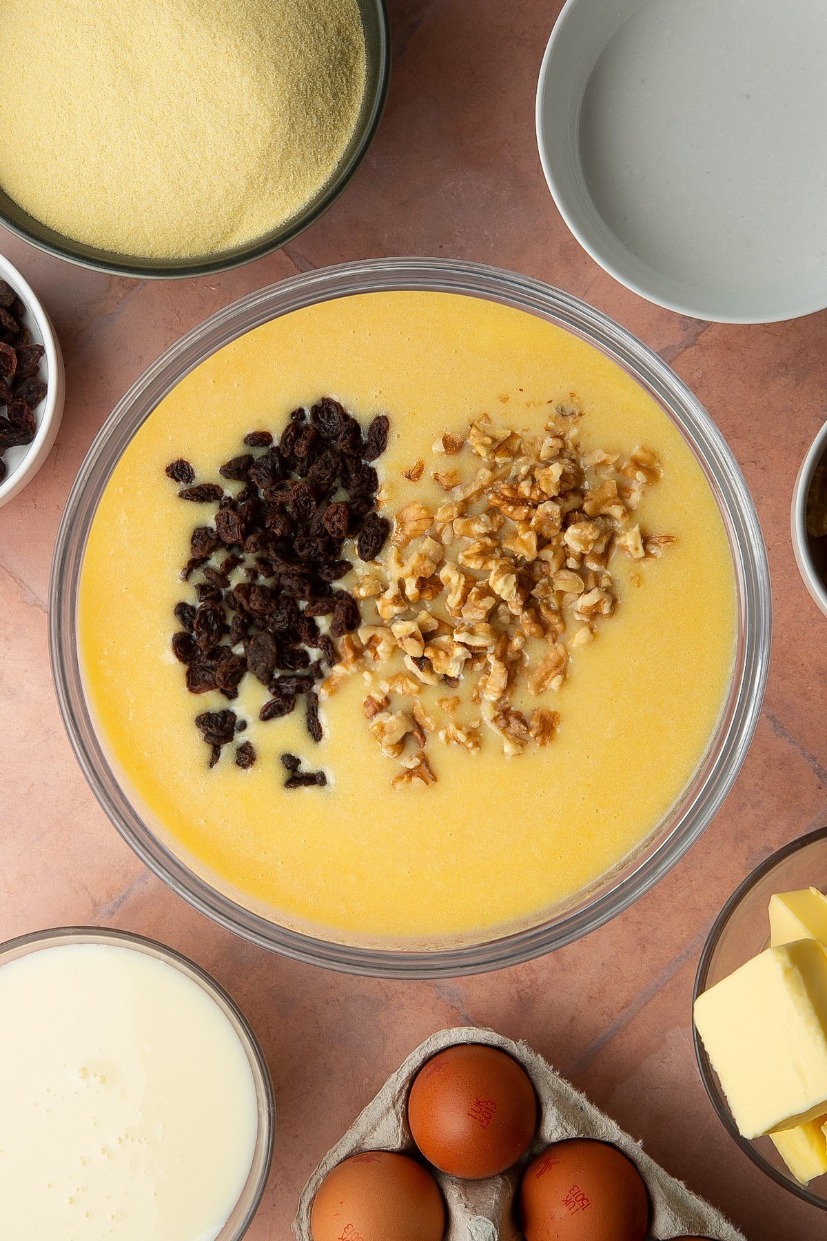 Golden semolina cake batter in a bowl with raisins and walnuts on top. Ingredients to make sanwin-makin surround the bowl.