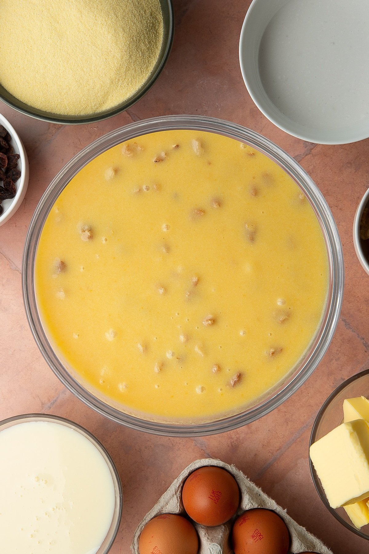 Golden semolina cake batter with raisins and walnuts in a bowl. Ingredients to make sanwin-makin surround the bowl.