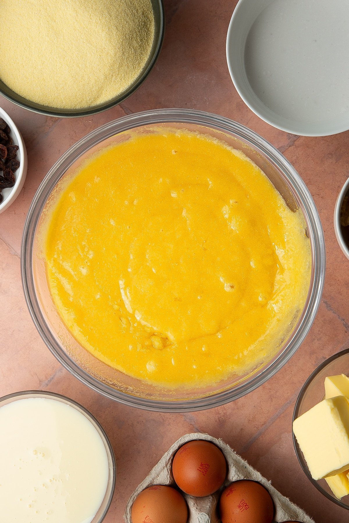 Semolina, sugar, eggs and melted butter mixed together in a bowl. Ingredients to make sanwin-makin surround the bowl.