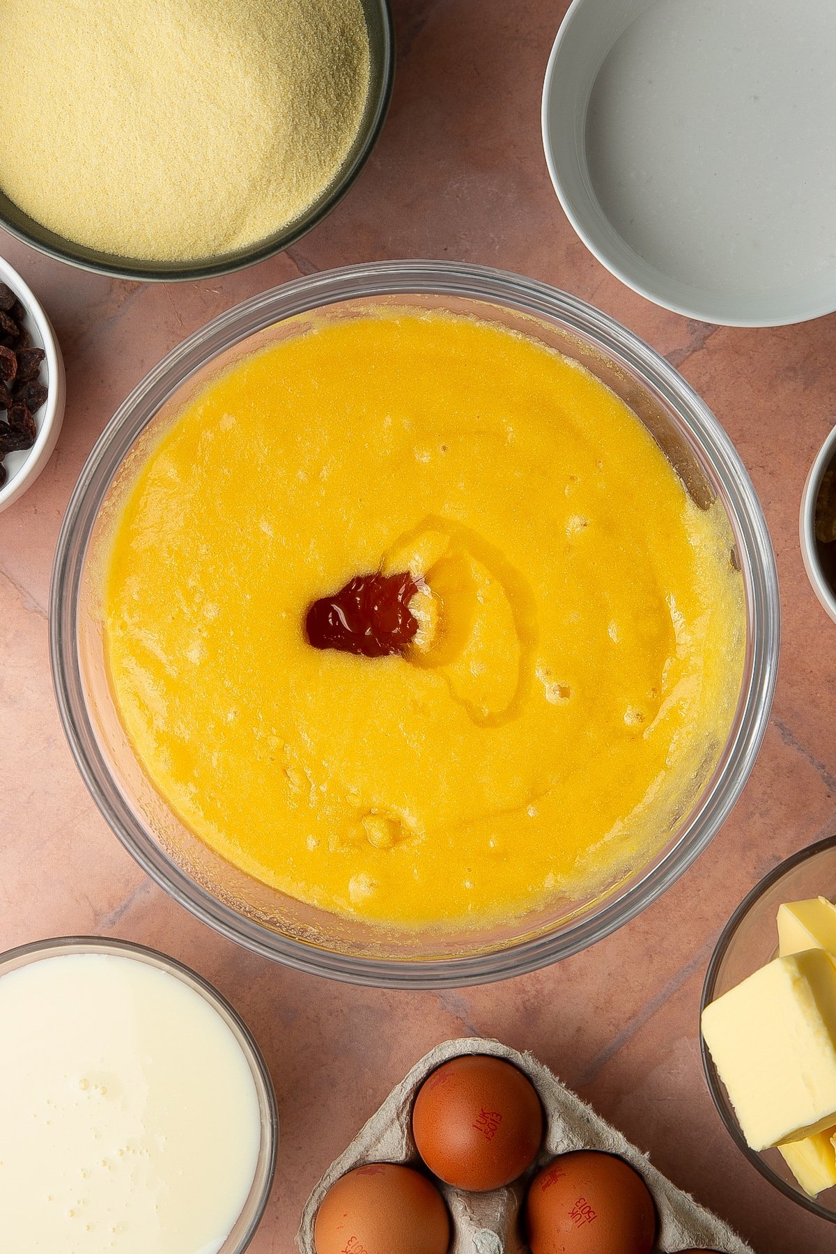 Semolina, sugar, eggs and melted butter mixed together in a bowl with golden syrup on top. Ingredients to make sanwin-makin surround the bowl.