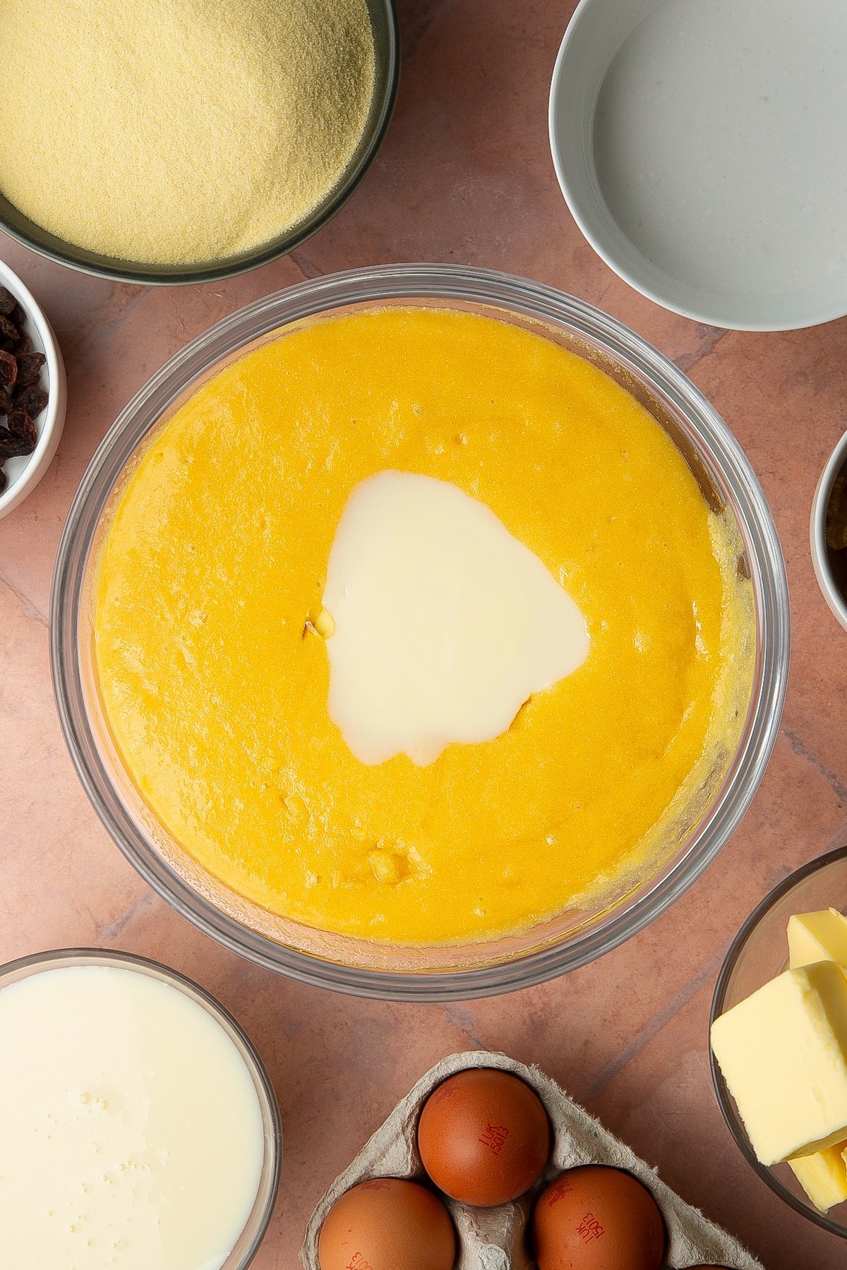 Semolina, sugar, eggs and melted butter and golden syrup mixed together in a bowl with condensed milk on top. Ingredients to make sanwin-makin surround the bowl.