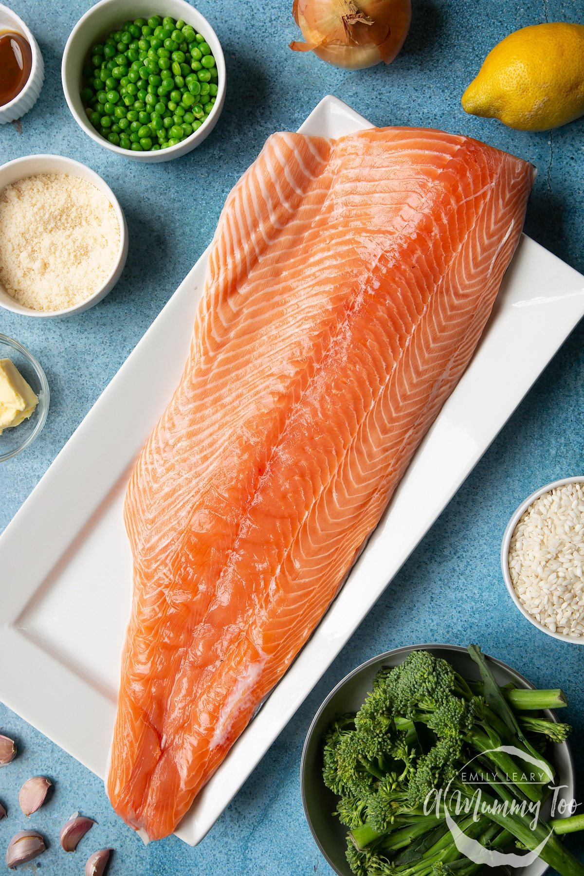 A side ot fresh Scottish salmon, skin-side down on a large, white, rectangular plate. Ingredients to make salmon risotto surround the plate.