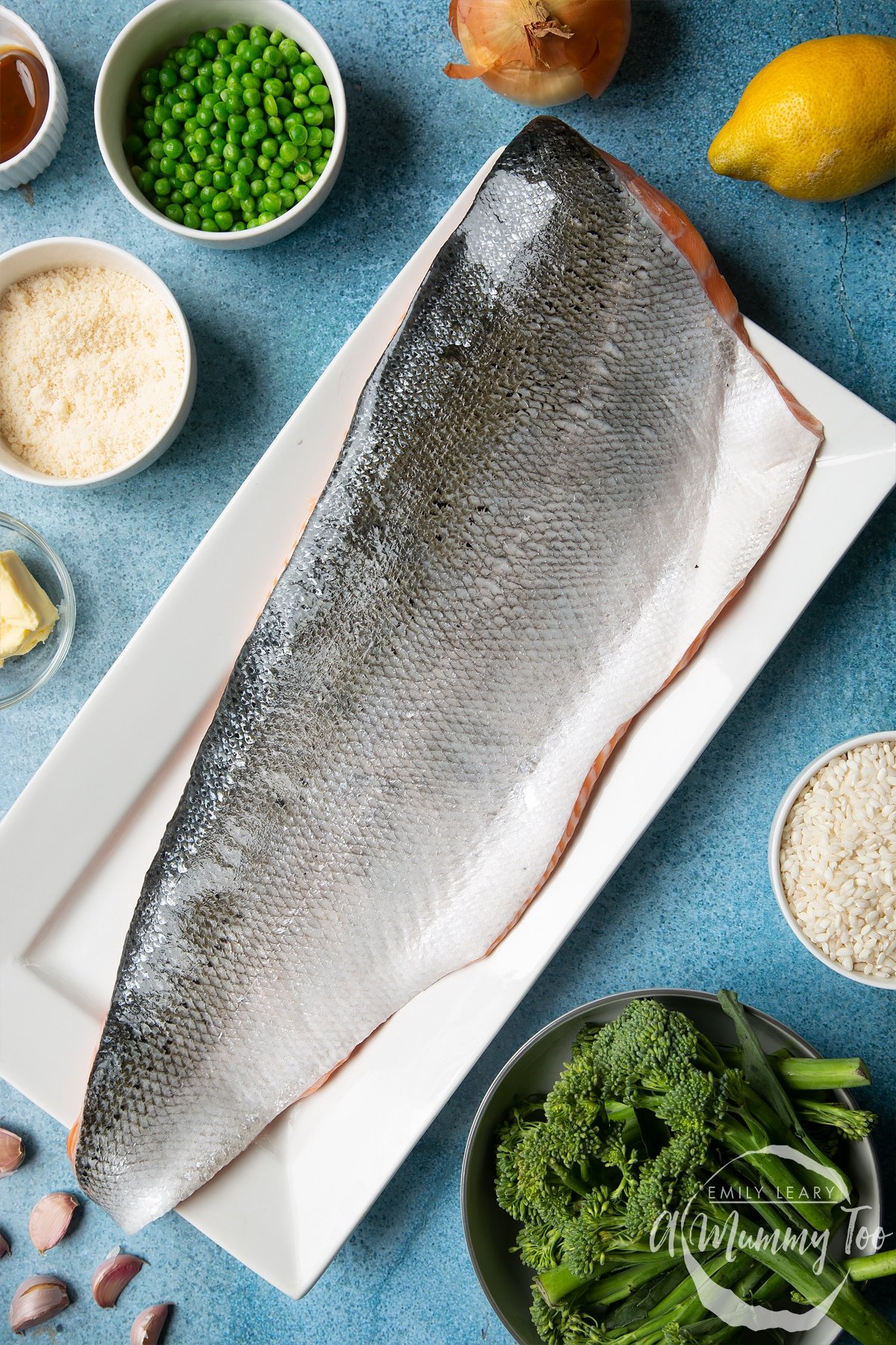 A side ot fresh Scottish salmon, skin-side up on a large, white, rectangular plate. Ingredients to make salmon risotto surround the plate.