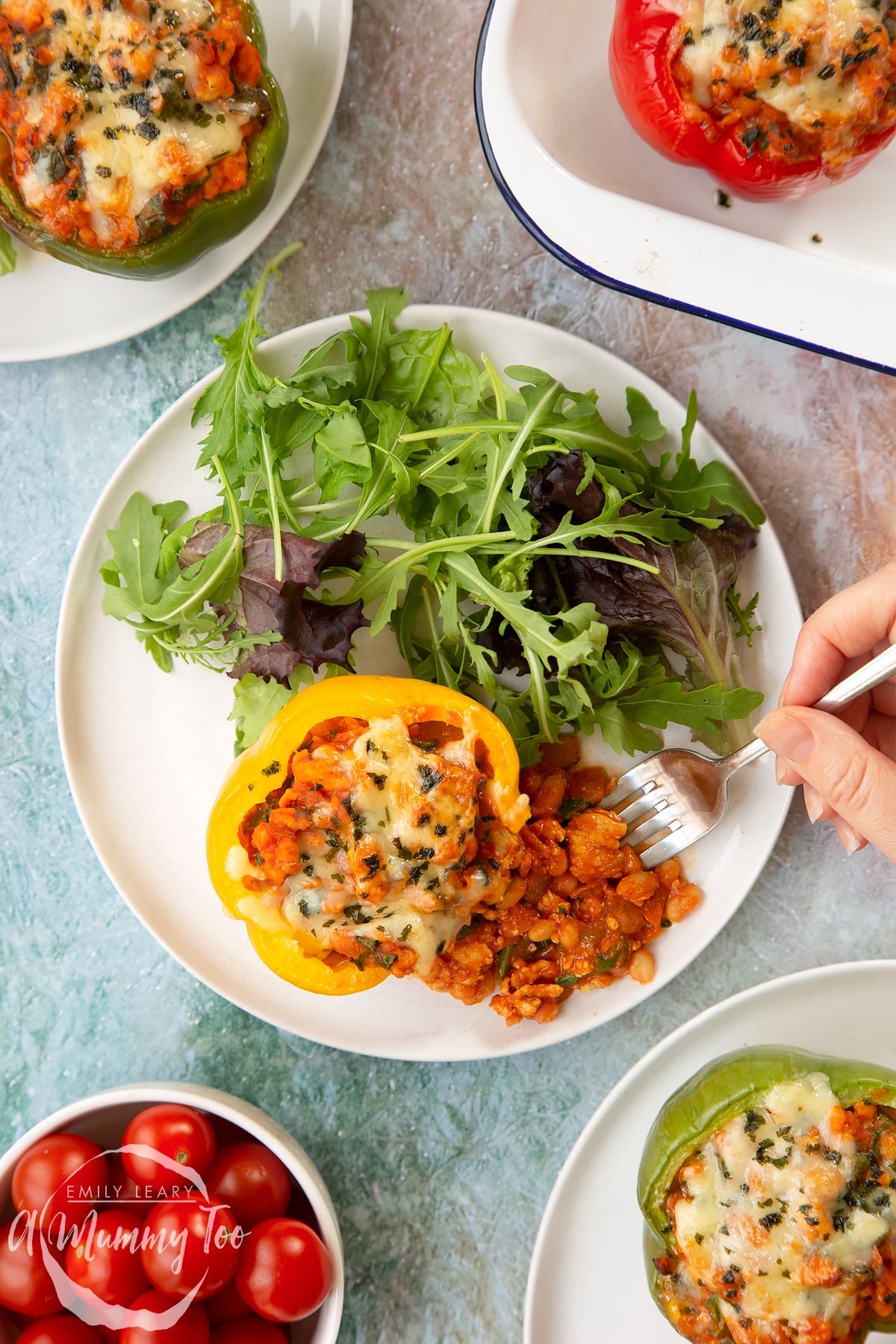 A yellow pepper stuffed with turkey mince, vegetables and baked beans with melted cheese on top. The filling spills onto the white plate and a fork delves into it. More baked bean stuffed peppers are shown to the edge of frame.