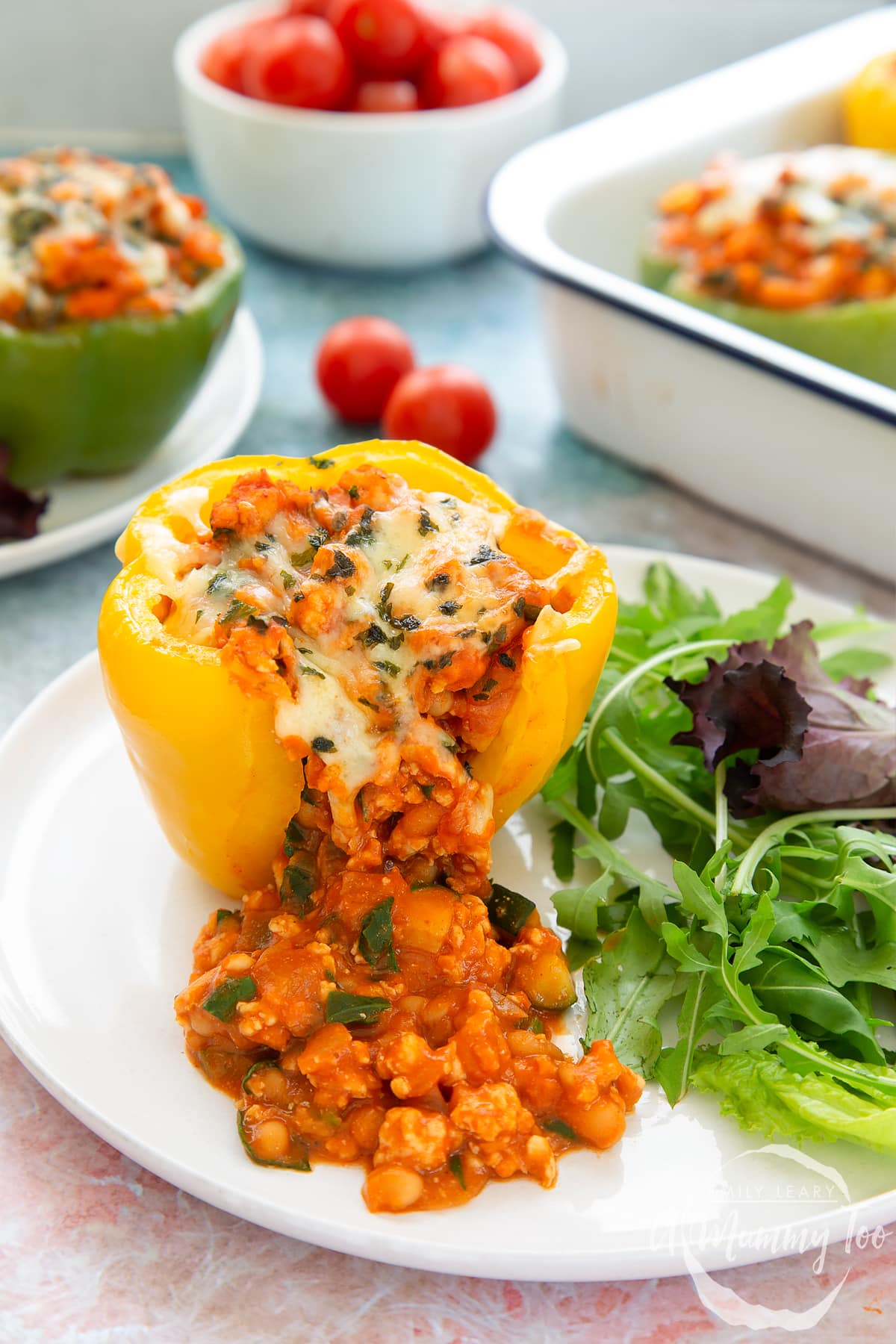 A yellow pepper stuffed with turkey mince, vegetables and baked beans with melted cheese on top. The filling spills onto the white plate. More baked bean stuffed peppers are shown in the background.