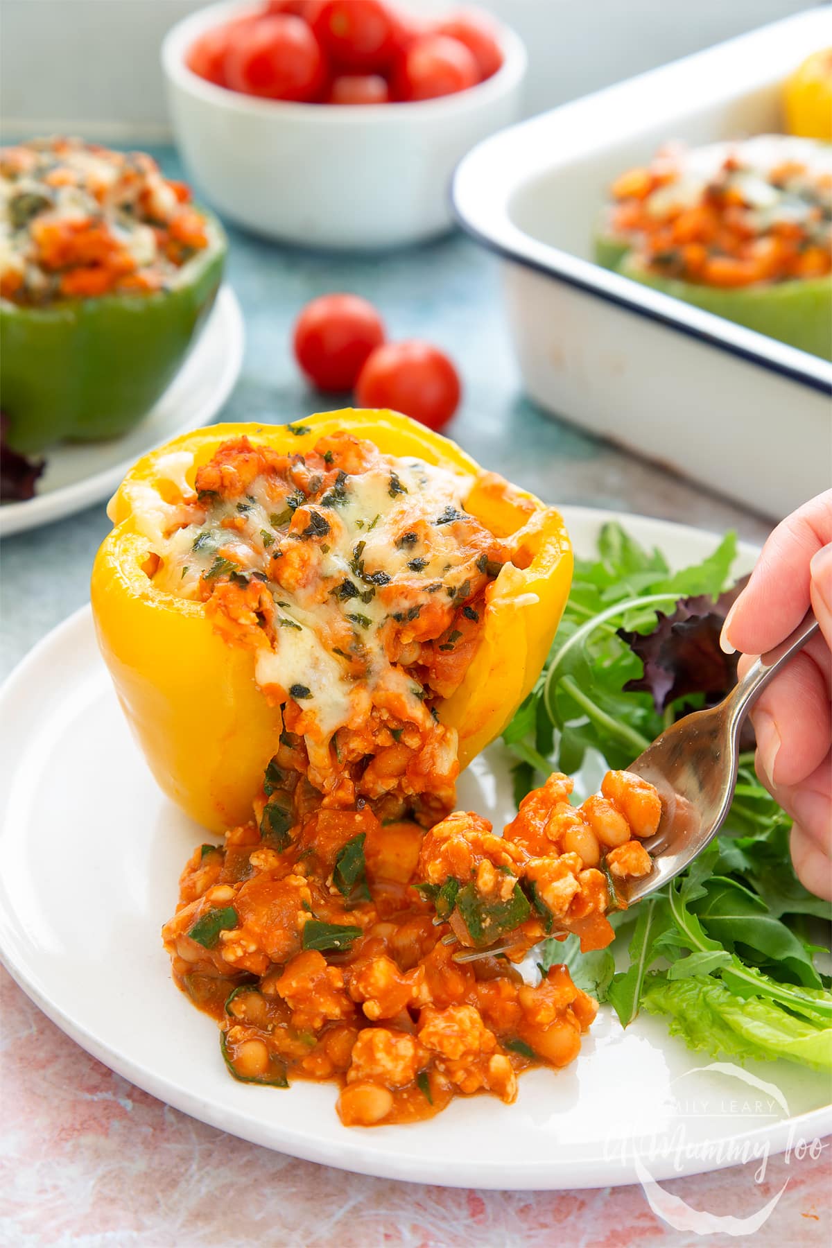A yellow pepper stuffed with turkey mince, vegetables and baked beans with melted cheese on top. The filling spills onto the white plate and a fork delves into it. More baked bean stuffed peppers are shown in the background.