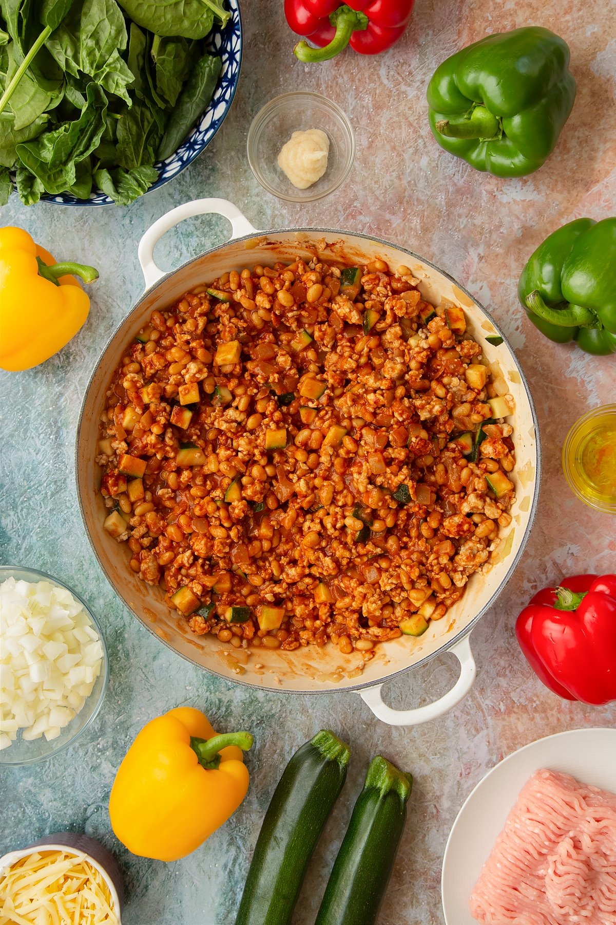 Onions, garlic, turkey mince, courgette, baked beans, turkey mince, tomato puree, cumin and smoked paprika in a large shallow pan. Ingredients to make baked bean stuffed peppers surround the pan.