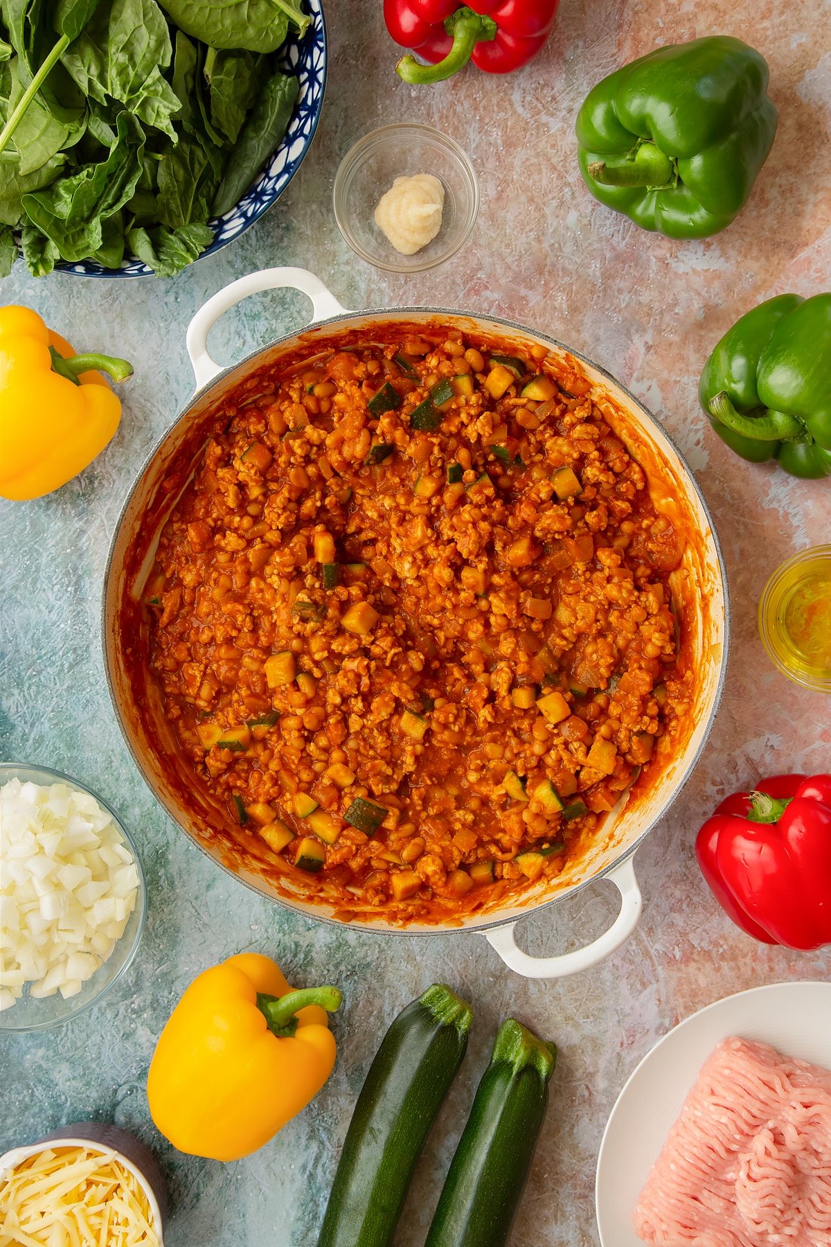 Vegetables, turkey mince and baked beans simmered in a spicy tomato sauce in a large shallow pan. Ingredients to make baked bean stuffed peppers surround the pan.