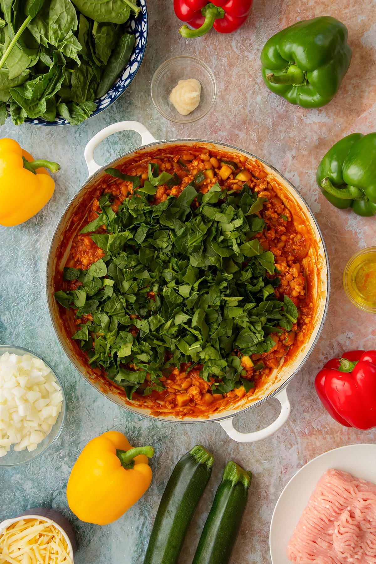 Vegetables, turkey mince and baked beans simmered in a spicy tomato sauce in a large shallow pan with spinach on top. Ingredients to make baked bean stuffed peppers surround the pan.