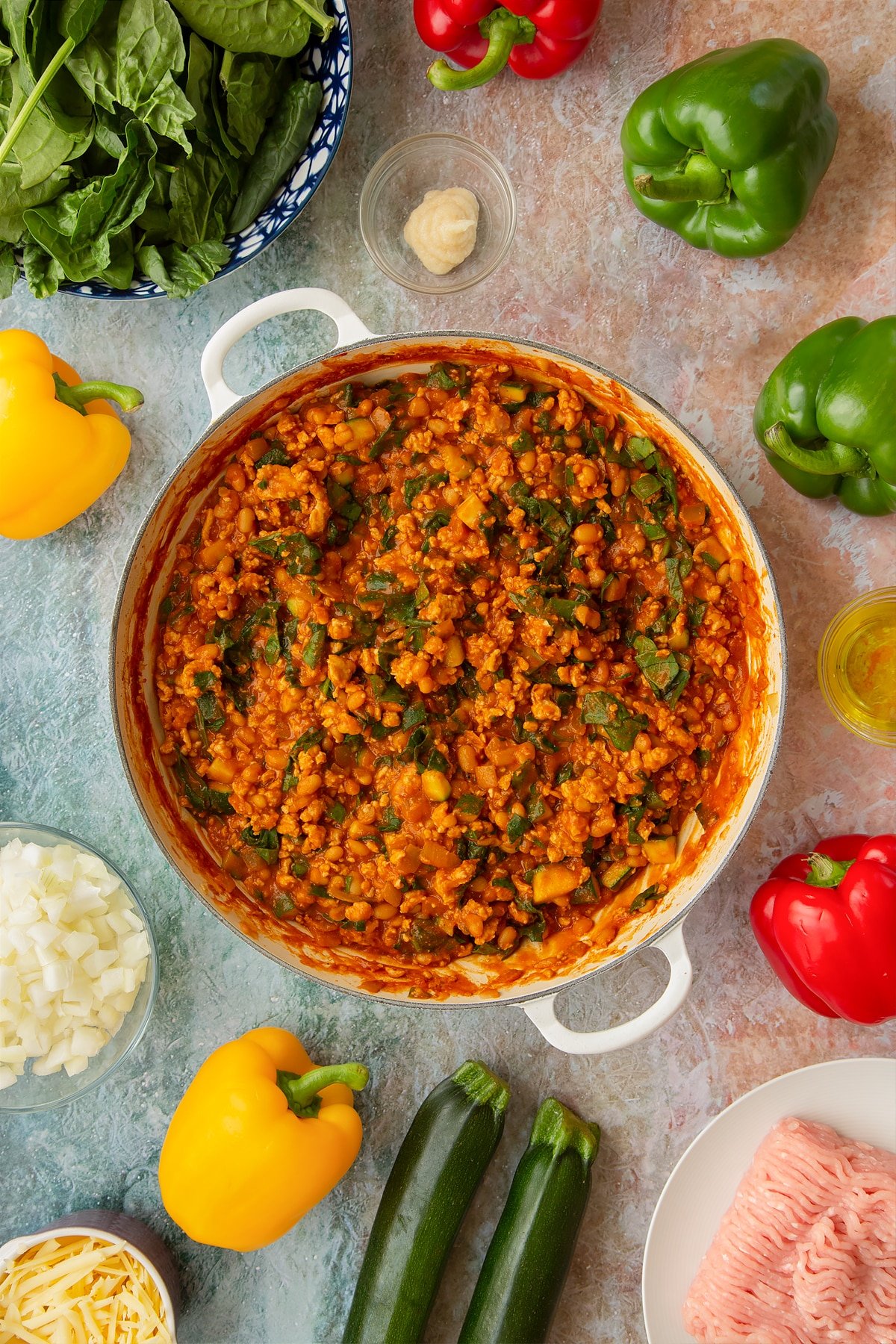 Vegetables, turkey mince, baked beans and wilted spinach in a spicy tomato sauce in a large shallow pan. Ingredients to make baked bean stuffed peppers surround the pan.