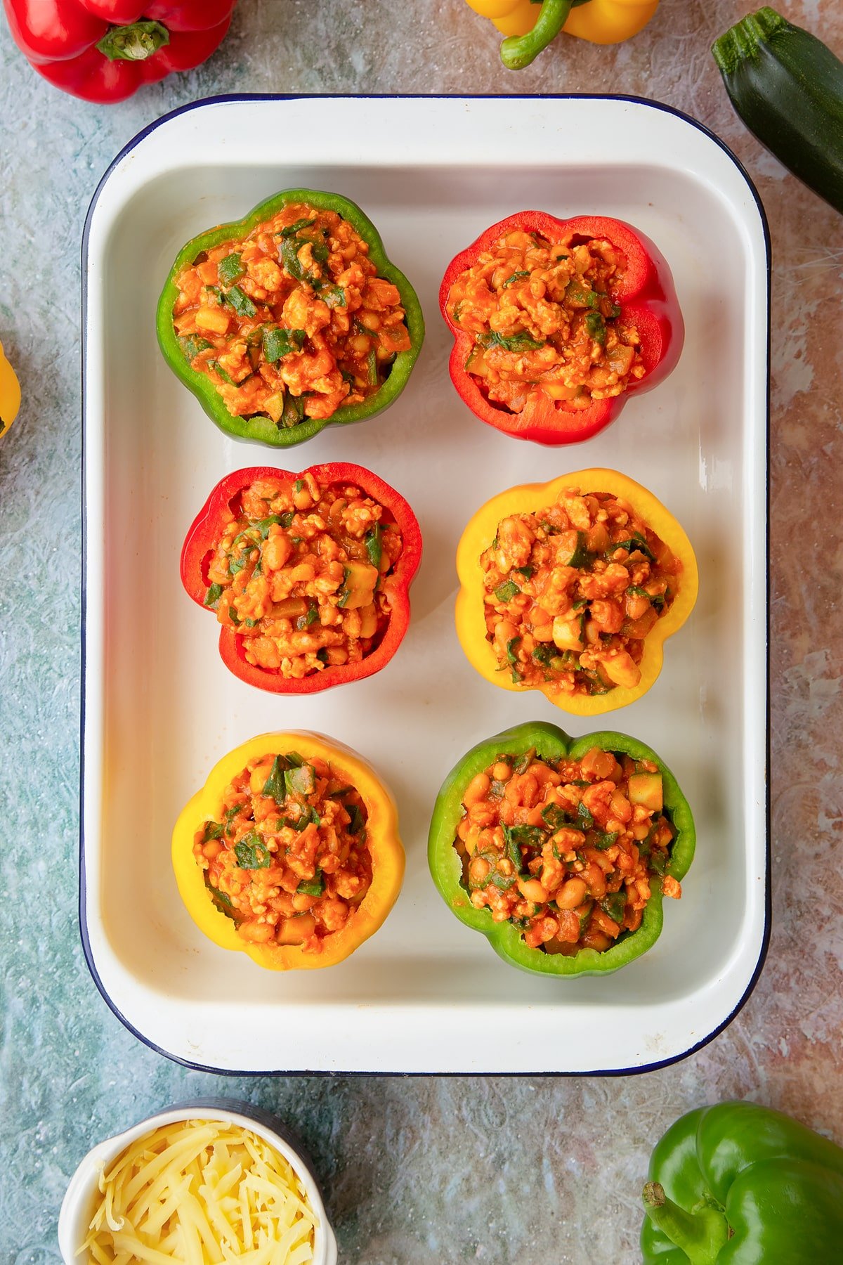 Six red, green and yellow peppers in a white roasting tray. The peppers are filled with the bean and turkey mix. Ingredients to make baked bean stuffed peppers surround the tray.