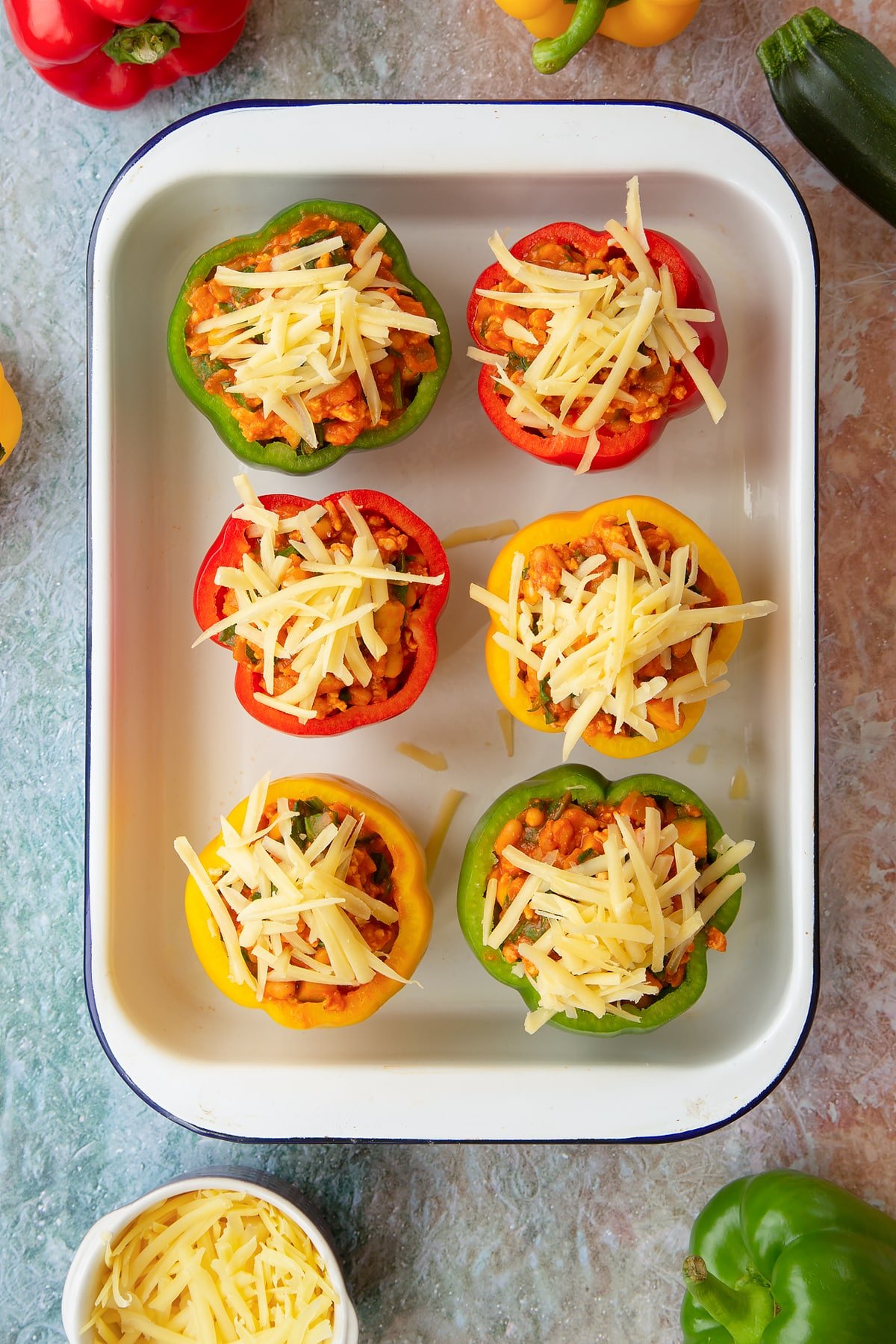 Six red, green and yellow peppers in a white roasting tray. The peppers are filled with the bean and turkey mix and topped with grated cheese. Ingredients to make baked bean stuffed peppers surround the tray.