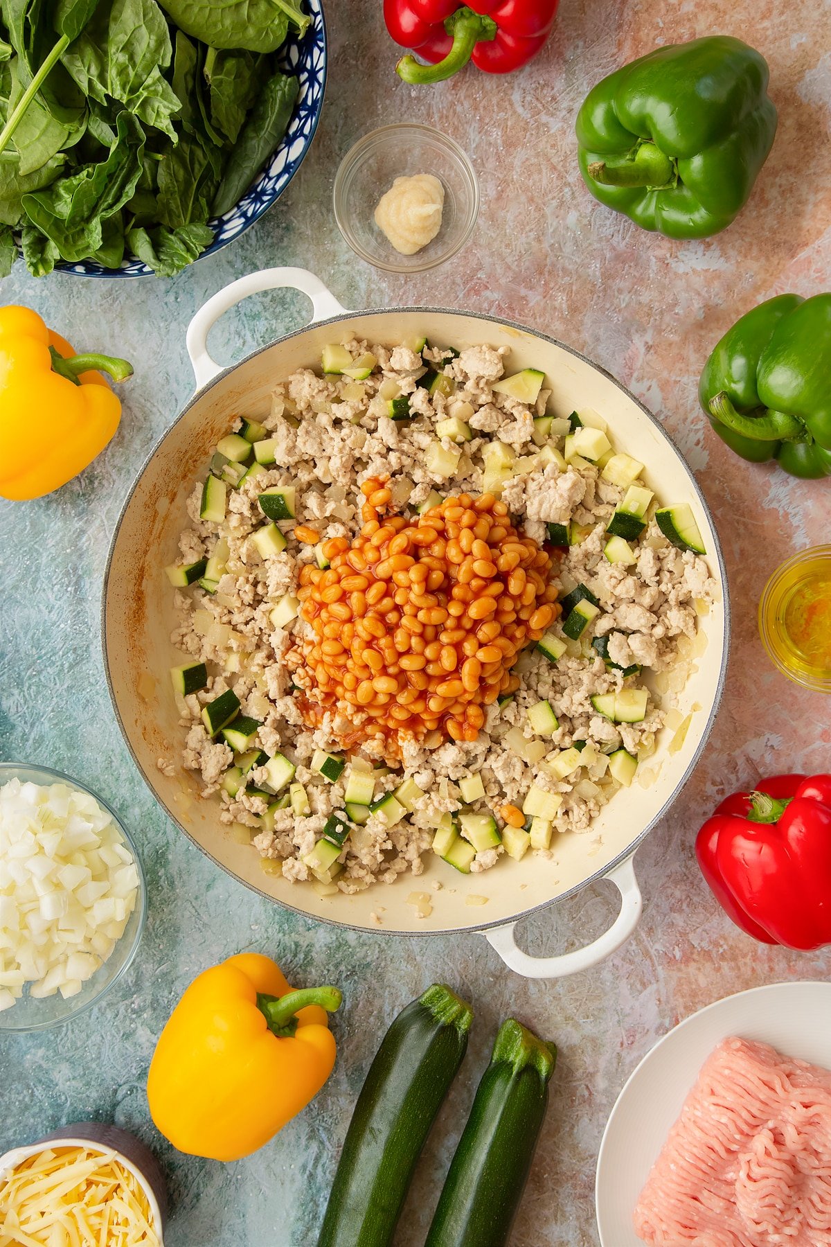 Lightly fried onions, garlic, turkey mince and courgette in a large shallow pan with baked beans on top. Ingredients to make baked bean stuffed peppers surround the pan.