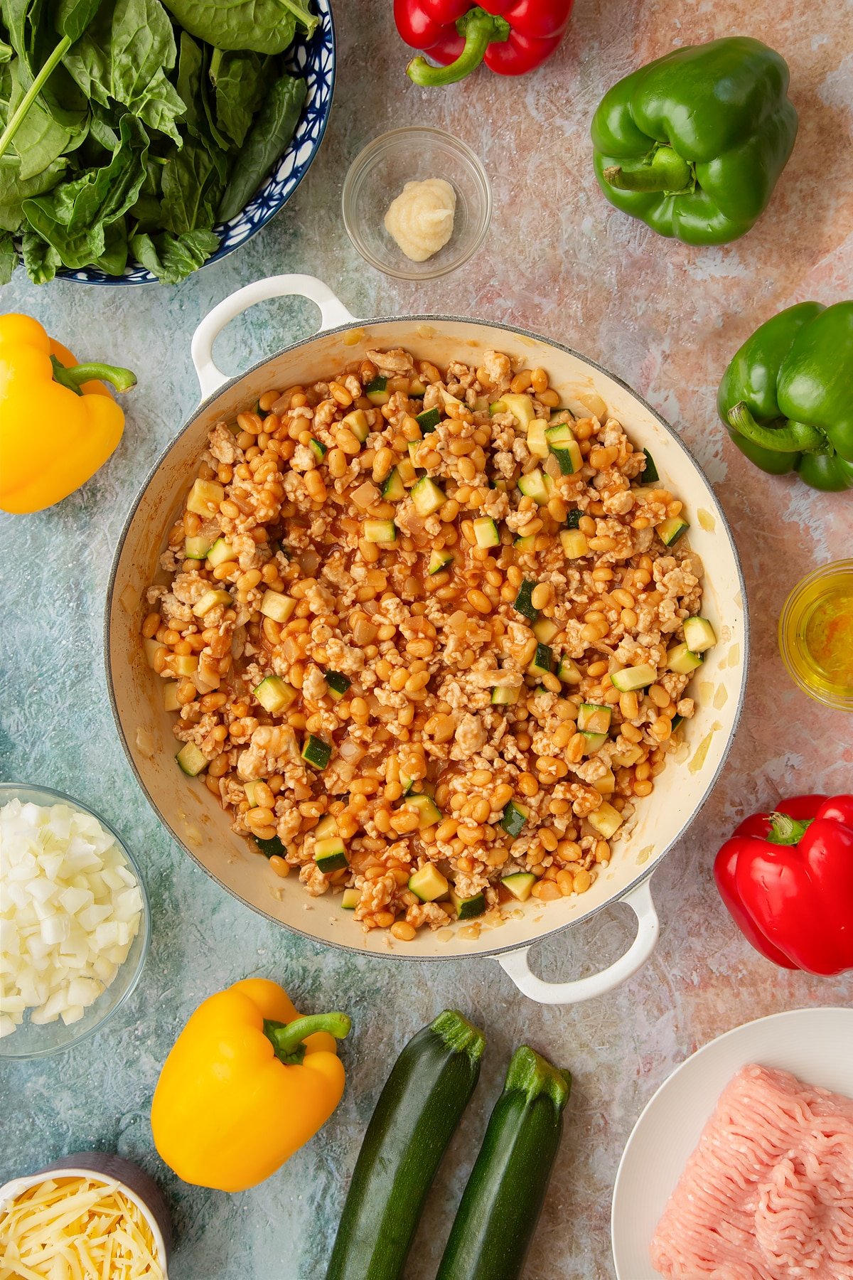 Onions, garlic, turkey mince, courgette, baked beans and turkey mince in a large shallow pan. Ingredients to make baked bean stuffed peppers surround the pan.