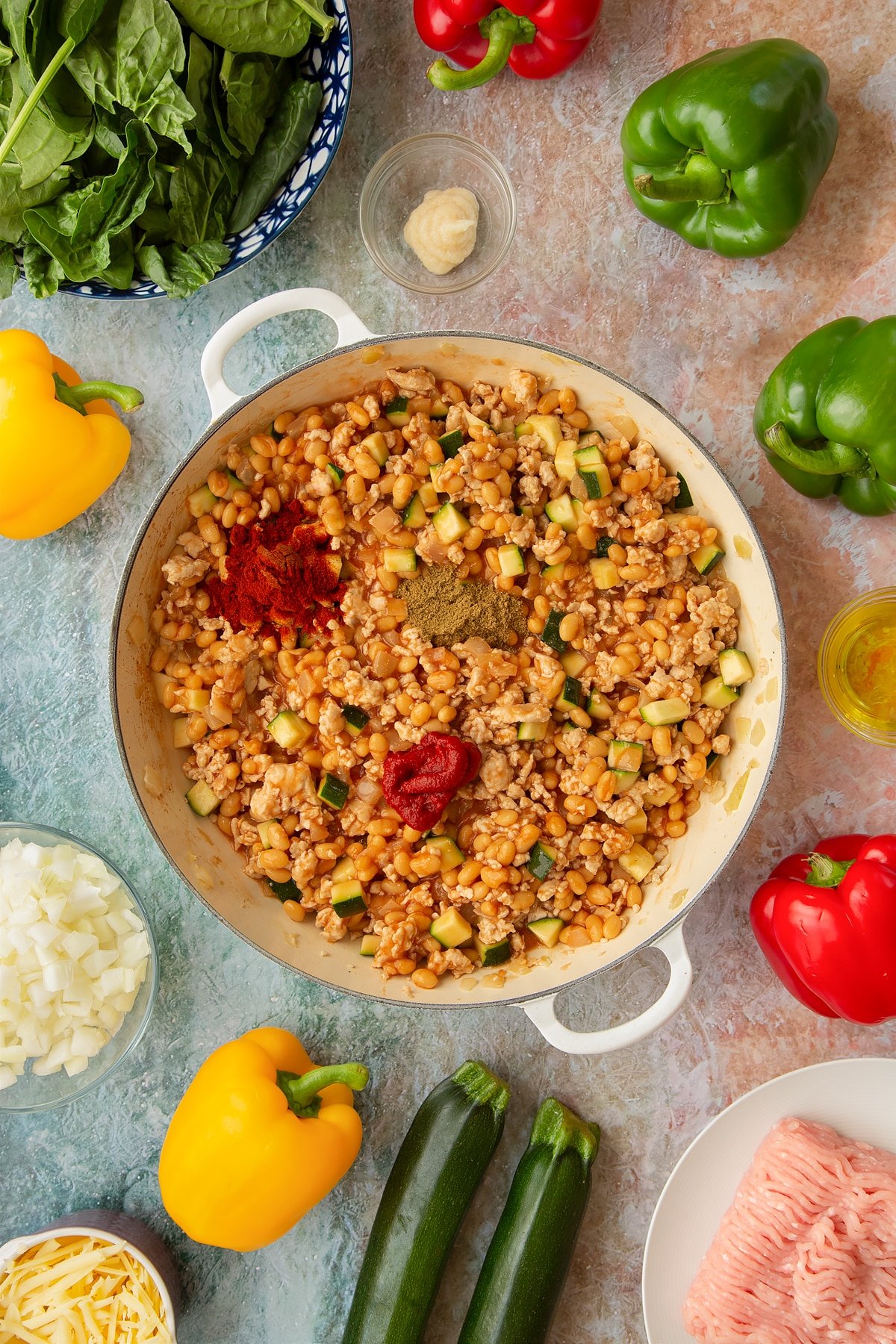 Onions, garlic, turkey mince, courgette, baked beans and turkey mince in a large shallow pan, with tomato puree, cumin and smoked paprika on top. Ingredients to make baked bean stuffed peppers surround the pan.