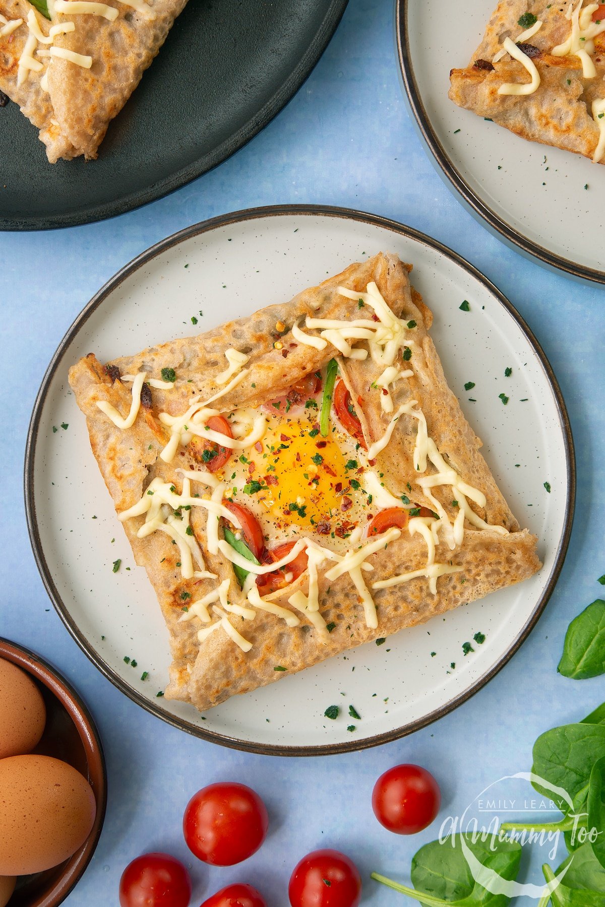 Overheat view of a buckwheat galette on plate with ingredients around it. The galette is filled with spinach, tomatoes, cheese and a lightly cooked egg. 