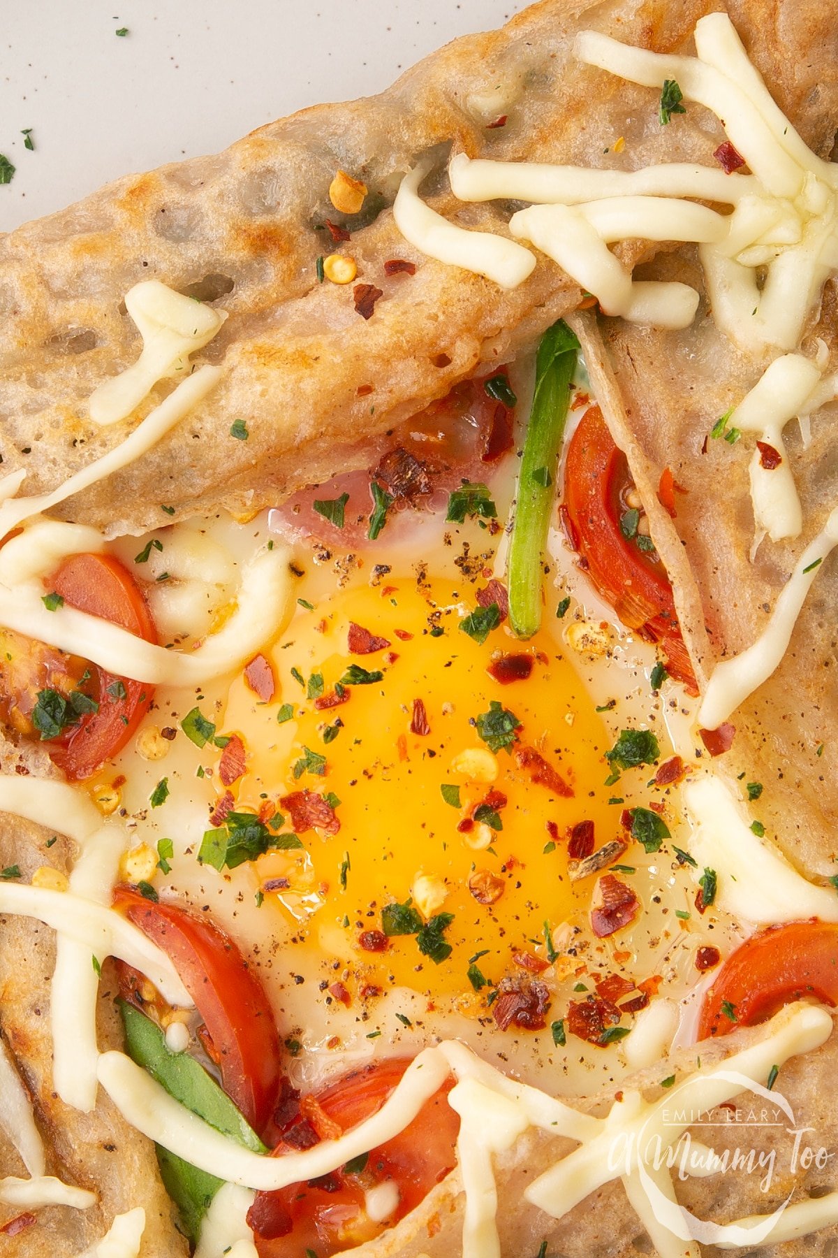 A buckwheat galette on plate, close up from above. The galette is filled with spinach, tomatoes, cheese and a lightly cooked egg. 