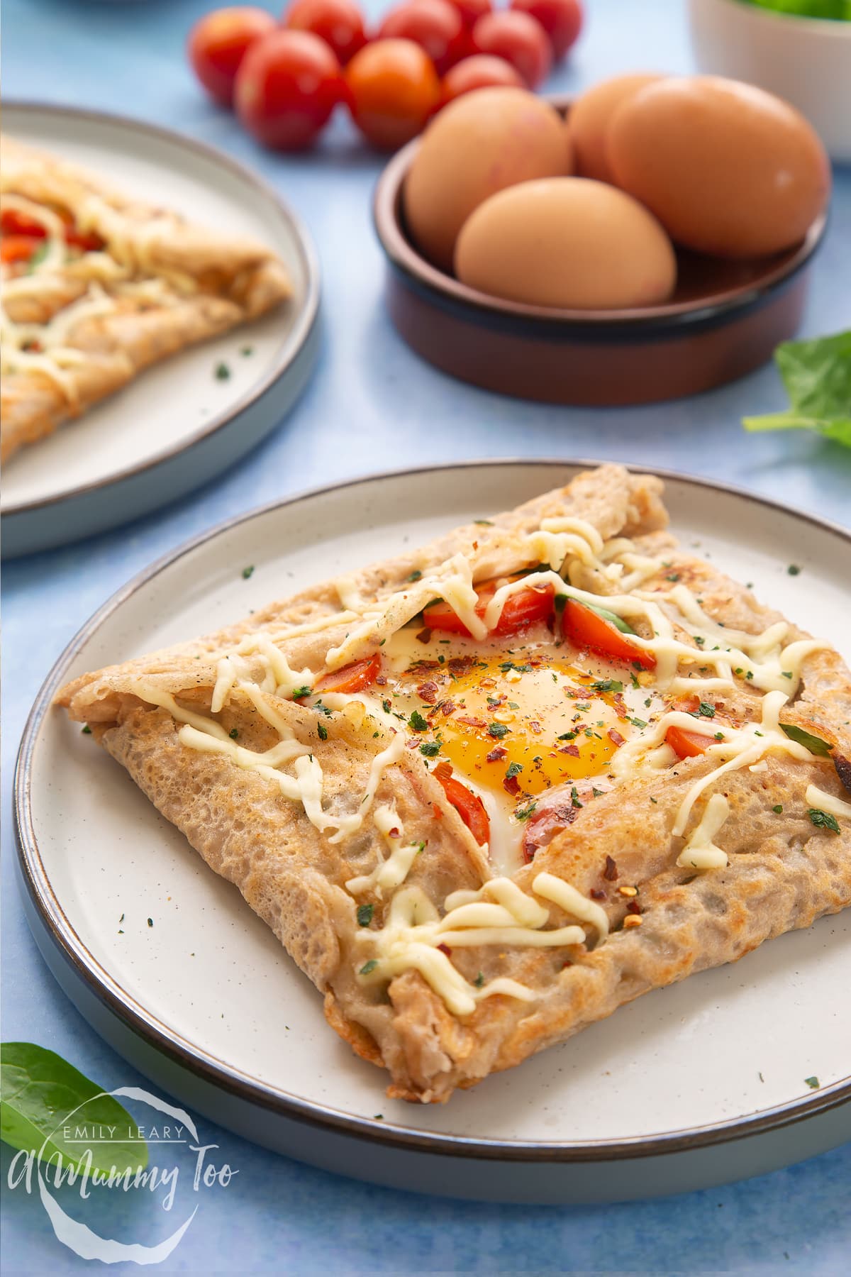 A buckwheat galette on plate with ingredients and another plate behind it. The galette is filled with spinach, tomatoes, cheese and a lightly cooked egg. 