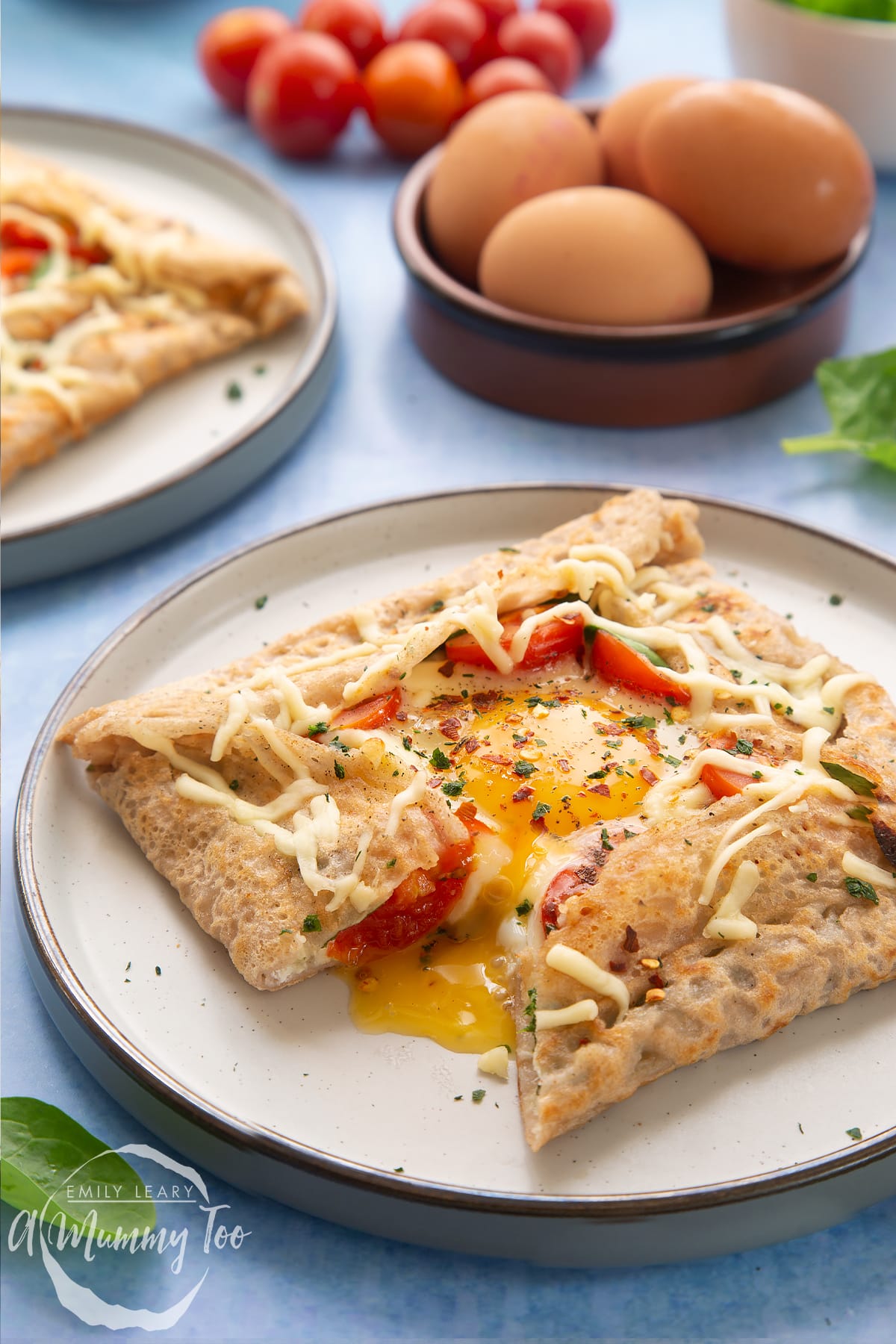 A buckwheat galette on plate with ingredients around it. The galette is filled with spinach, tomatoes, cheese and a lightly cooked egg. The galette has been cut to reveal the custardy yolk.