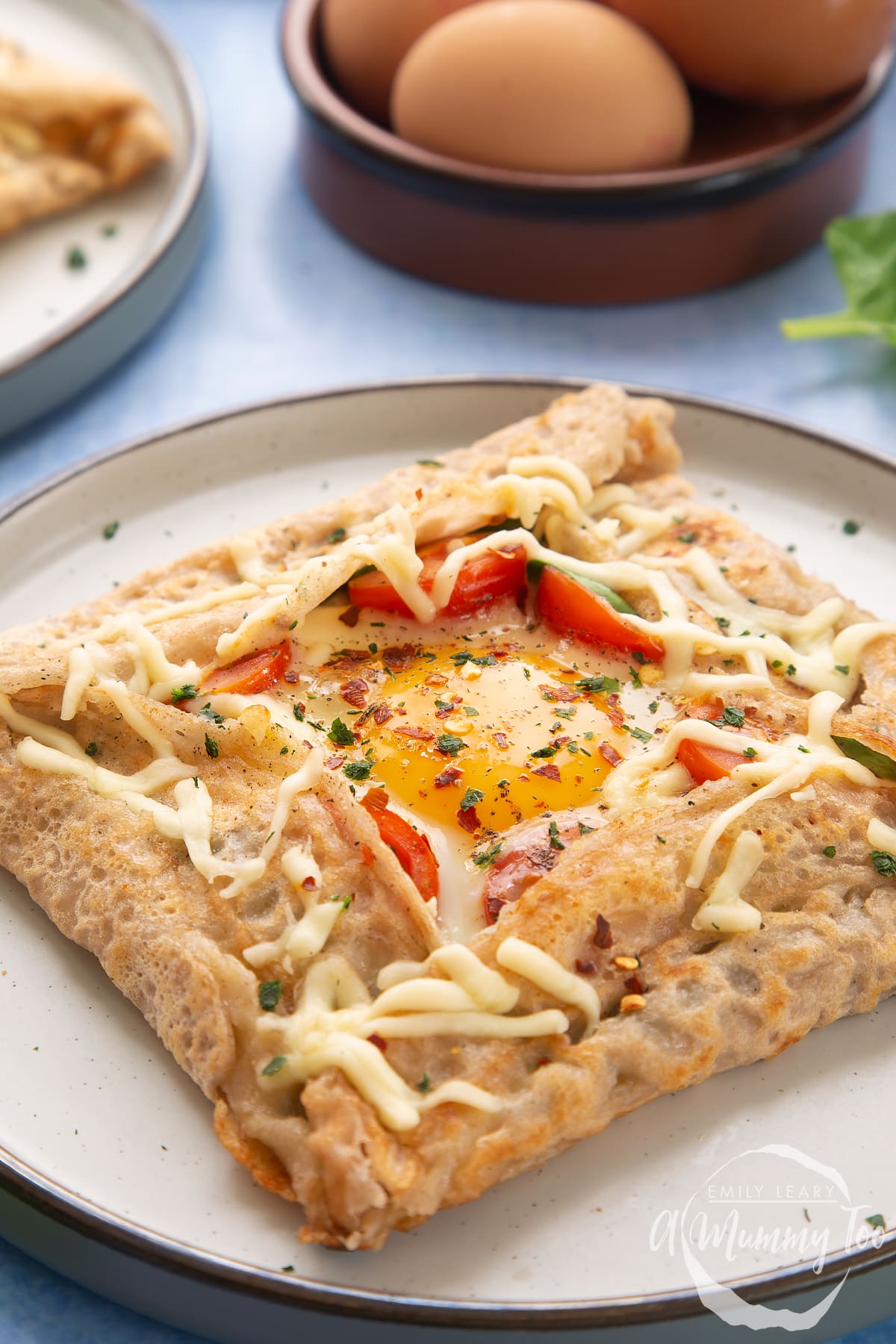 A buckwheat galette on plate with a dish of eggs behind it. The galette is filled with spinach, tomatoes, cheese and a lightly cooked egg. 