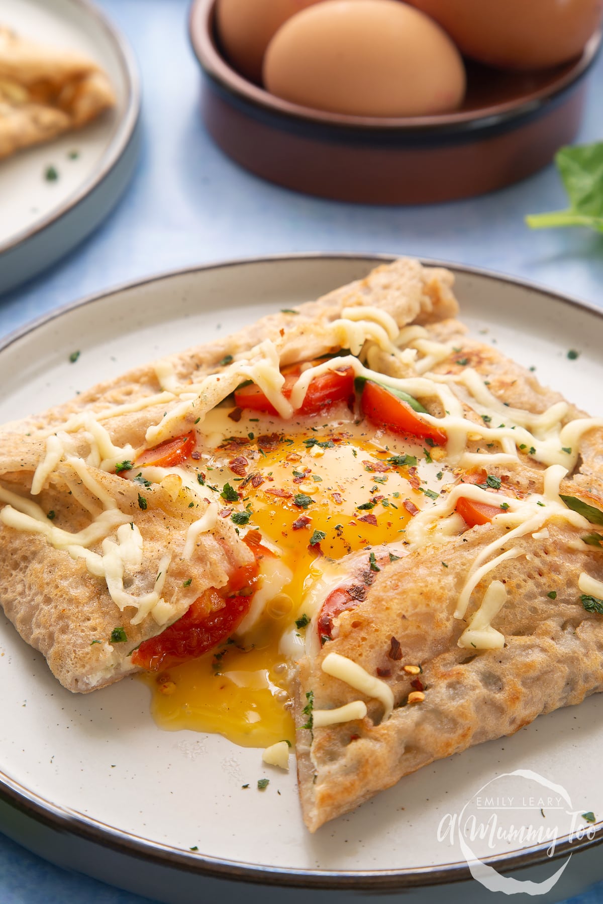 A buckwheat galette on plate with ingredients behind it. The galette is filled with spinach, tomatoes, cheese and a lightly cooked egg. 