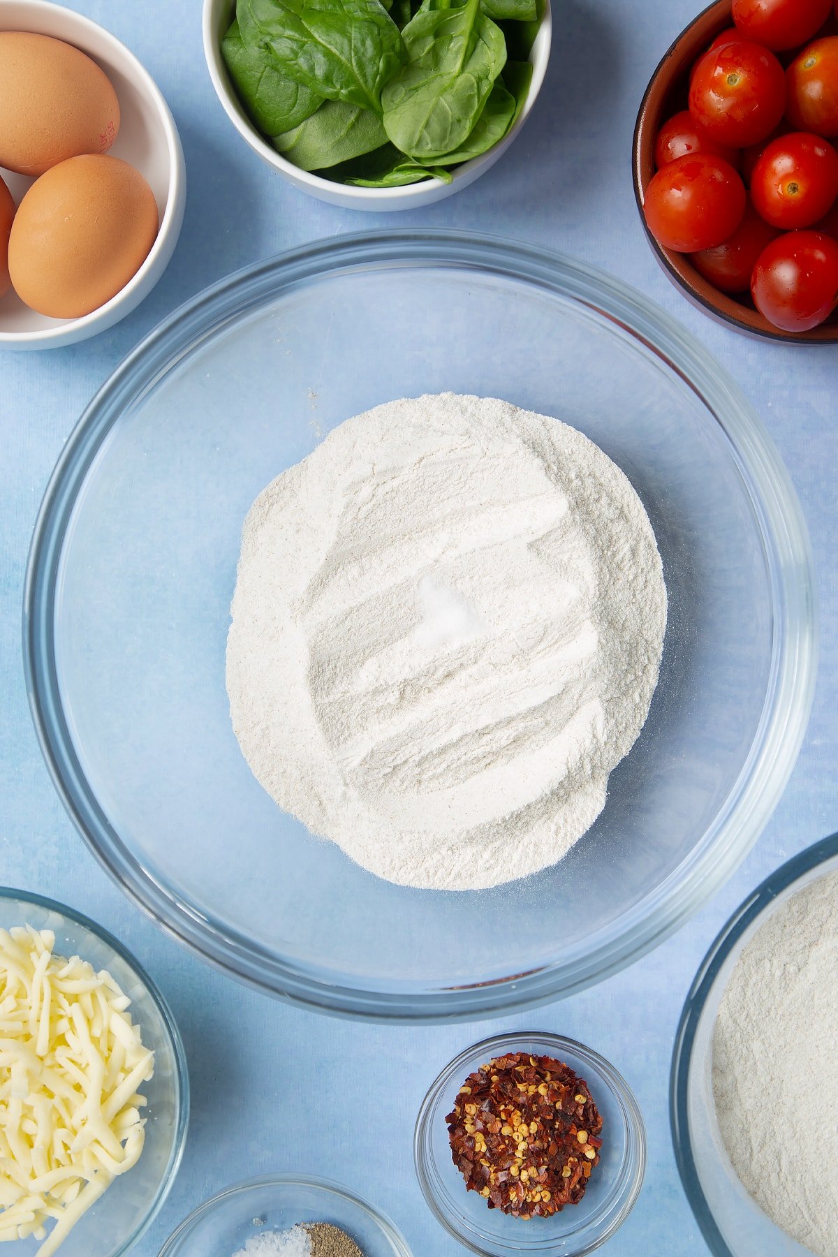 Buckwheat flour in a glass mixing bowl. Ingredients to make buckwheat galettes surround the bowl.