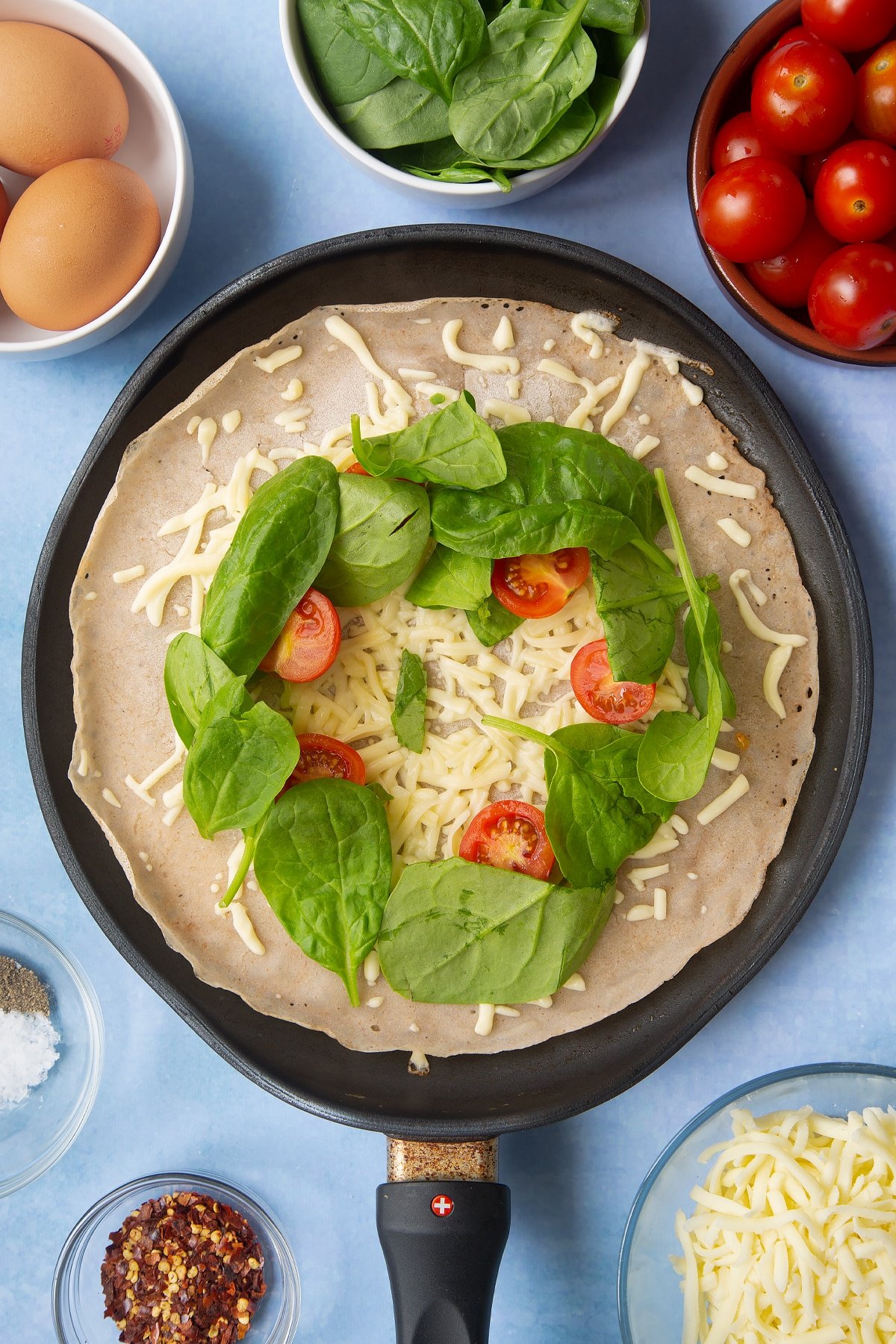 Buckwheat crepe topped with grated cheese, cherry tomatoes and spinach in a crepe pan. Ingredients to make buckwheat galettes surround the pan.