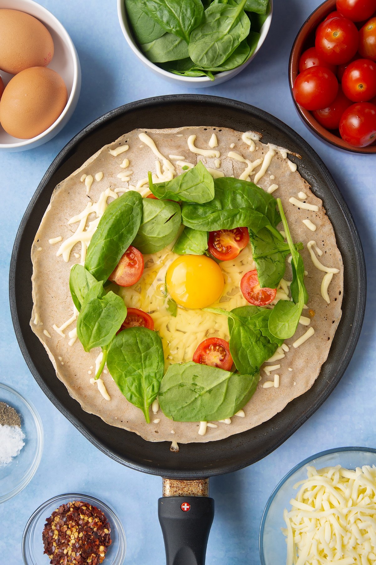 Buckwheat crepe topped with grated cheese, cherry tomatoes and spinach in a crepe pan. An egg has been cracked into the centre. Ingredients to make buckwheat galettes surround the pan.