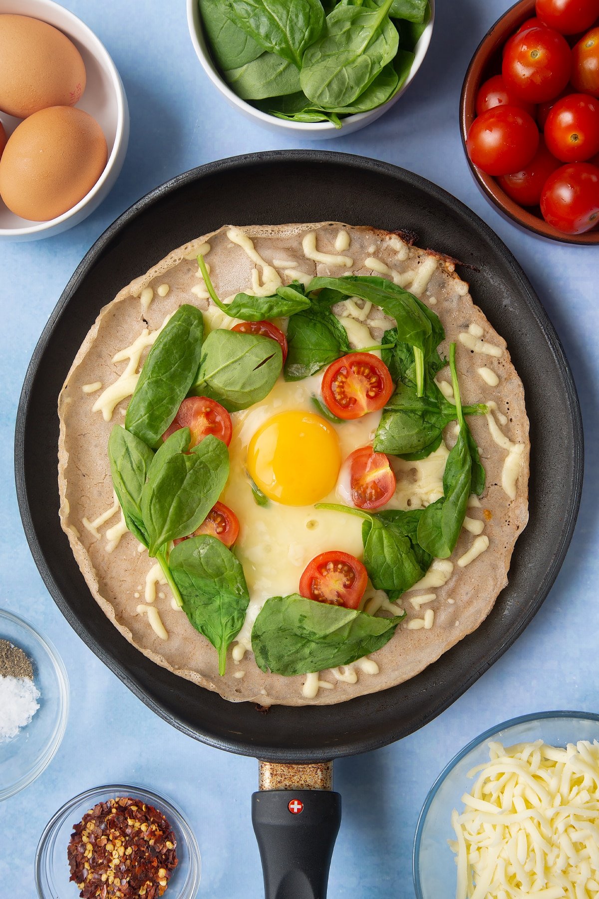 Buckwheat crepe topped with grated cheese, cherry tomatoes and spinach in a crepe pan. An egg is cooked in the centre. Ingredients to make buckwheat galettes surround the pan.