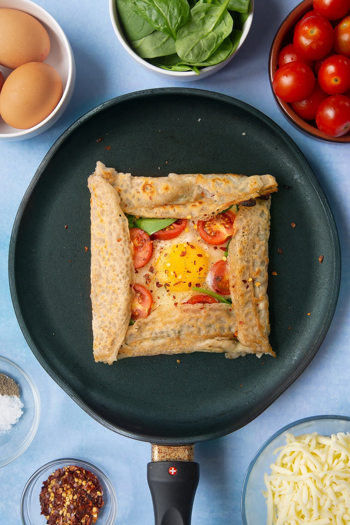Folded buckwheat galette in a crepe pan, scattered with chilli flakes. Ingredients to make buckwheat galettes surround the pan.