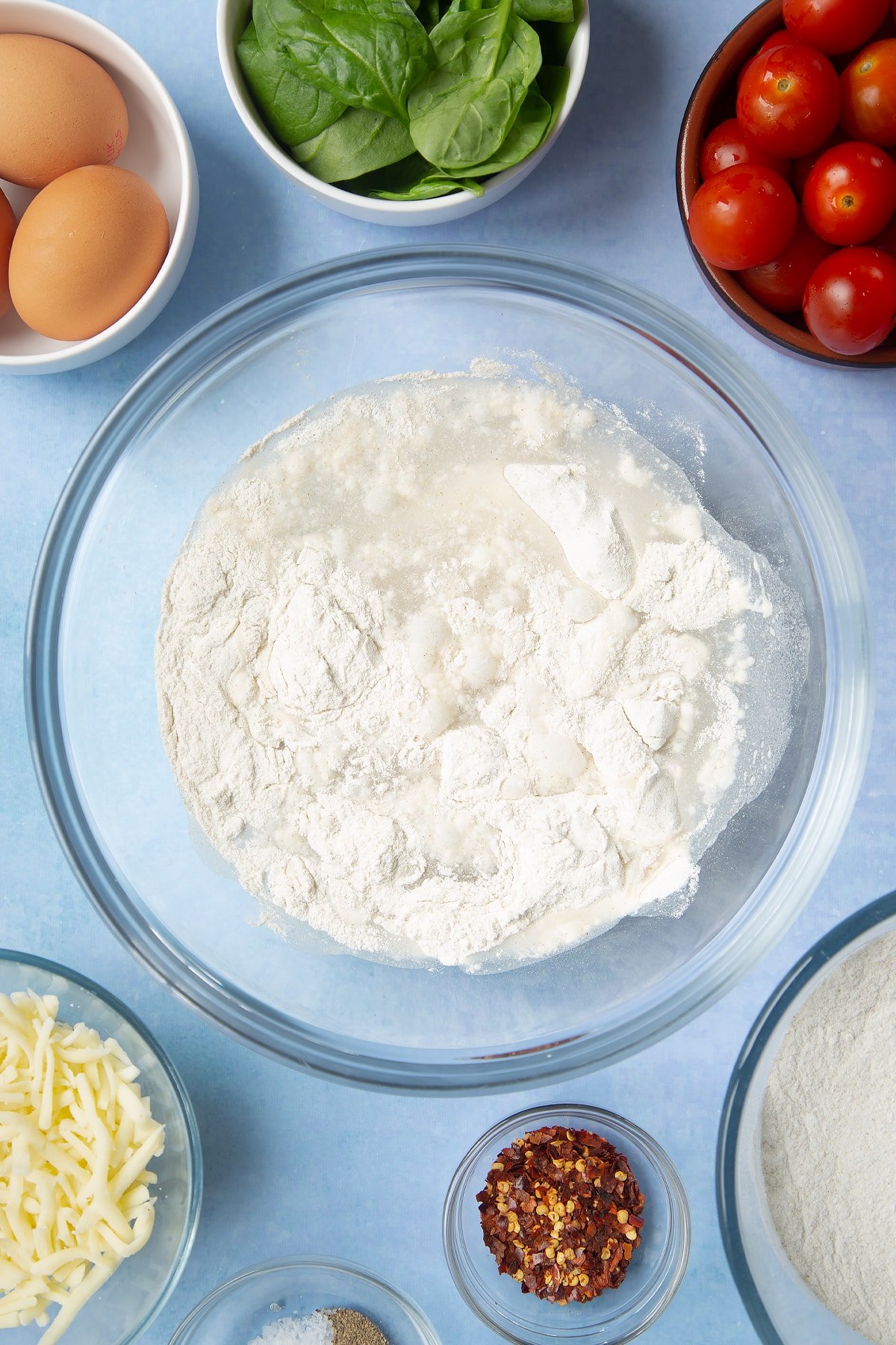 Buckwheat flour and water in a glass mixing bowl. Ingredients to make buckwheat galettes surround the bowl.