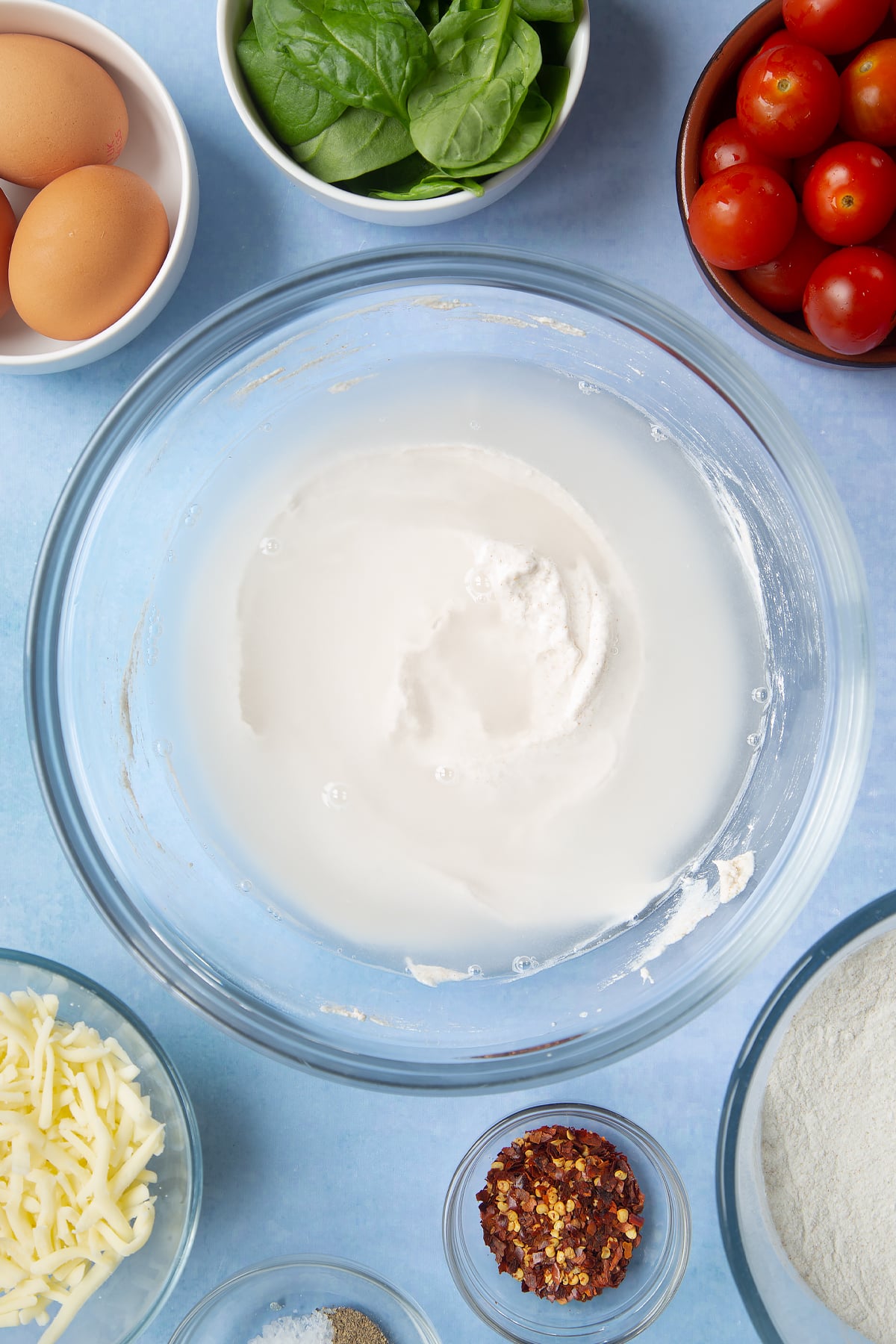 Buckwheat flour and water paste, with extra water on top in a glass mixing bowl. Ingredients to make buckwheat galettes surround the bowl.