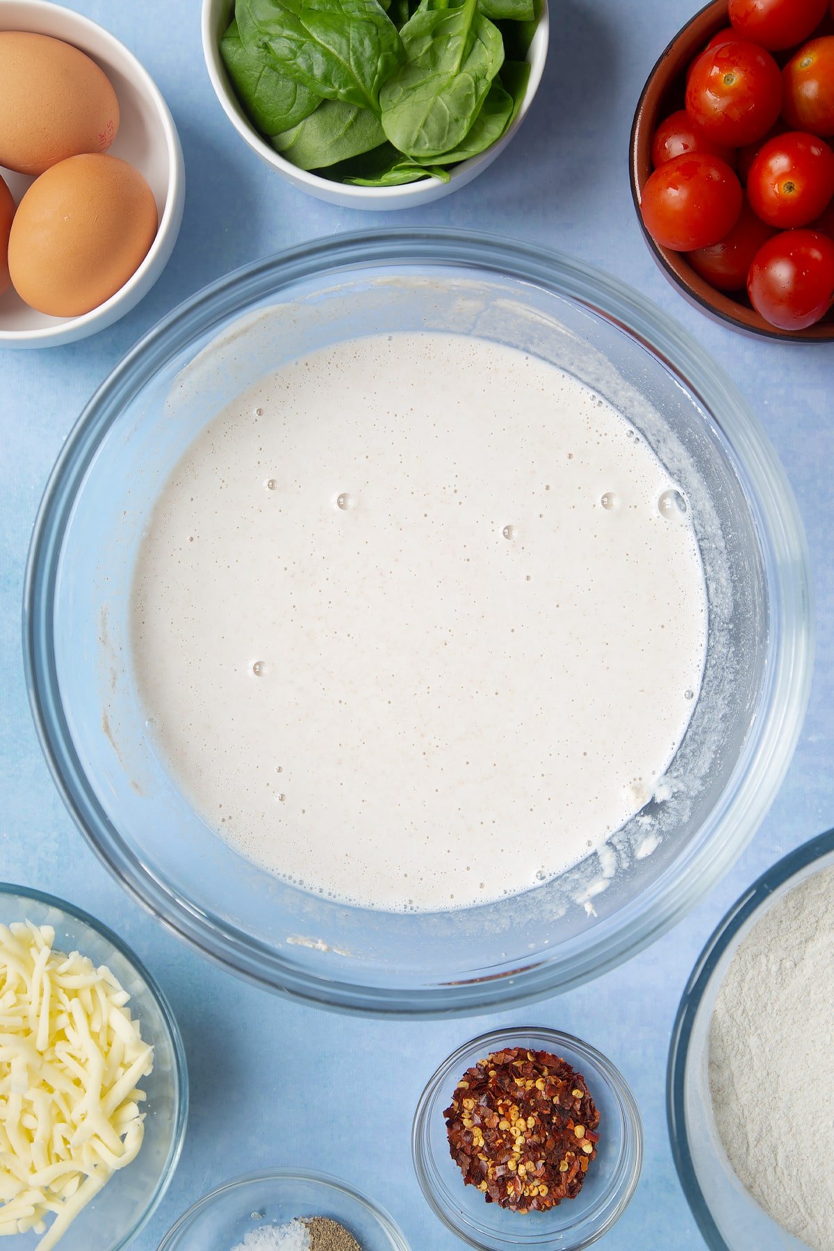 Buckwheat crepe batter in a glass mixing bowl. Ingredients to make buckwheat galettes surround the bowl.