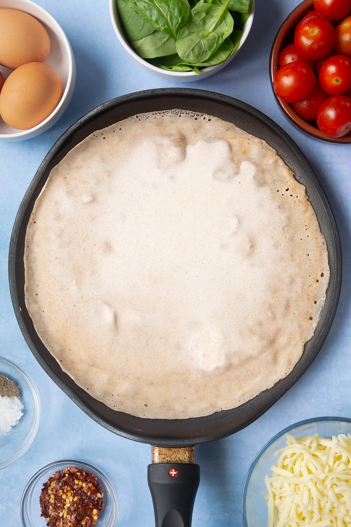 Buckwheat crepe batter in a crepe pan. Ingredients to make buckwheat galettes surround the pan.