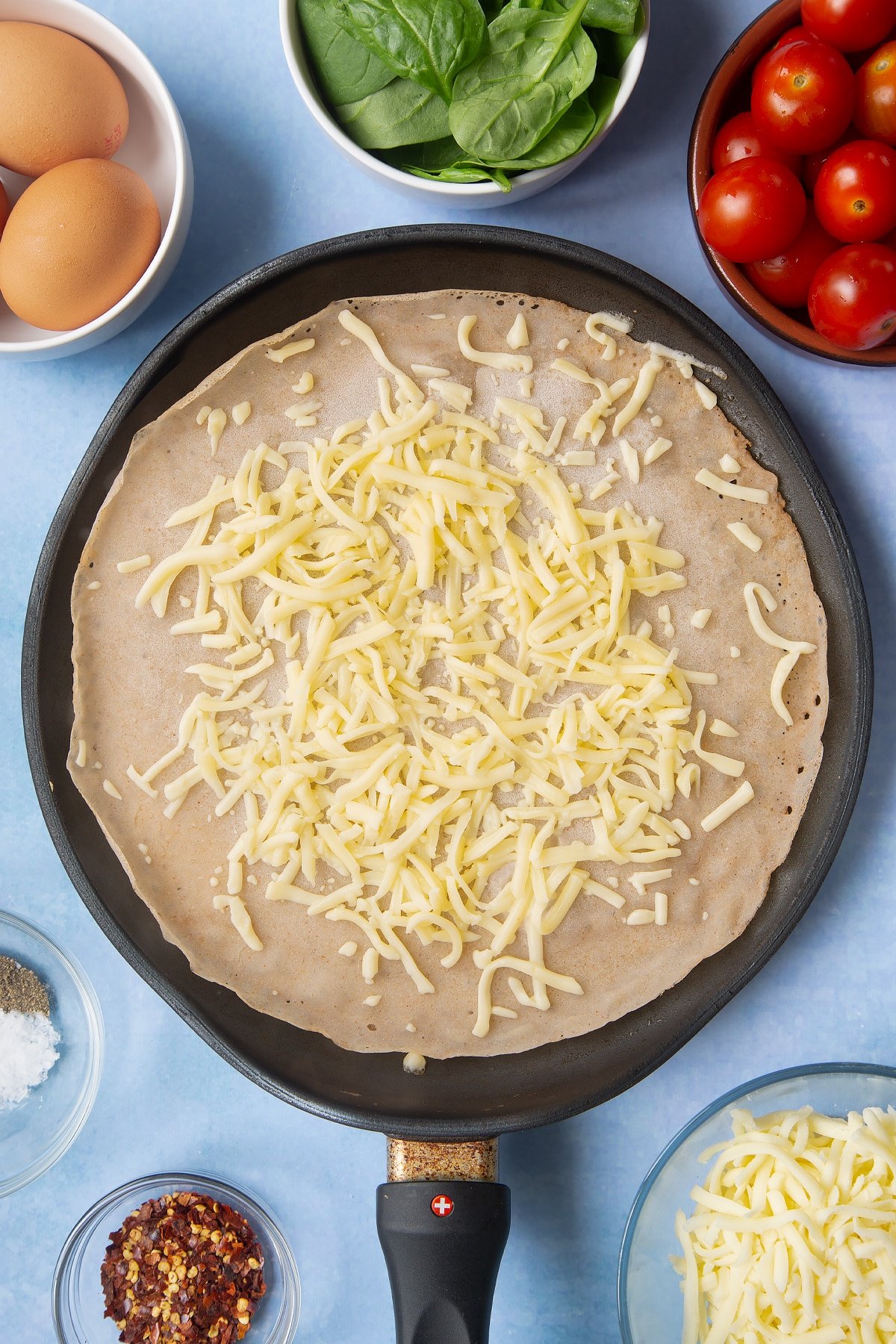 Buckwheat crepe topped with grated cheese in a crepe pan. Ingredients to make buckwheat galettes surround the pan.