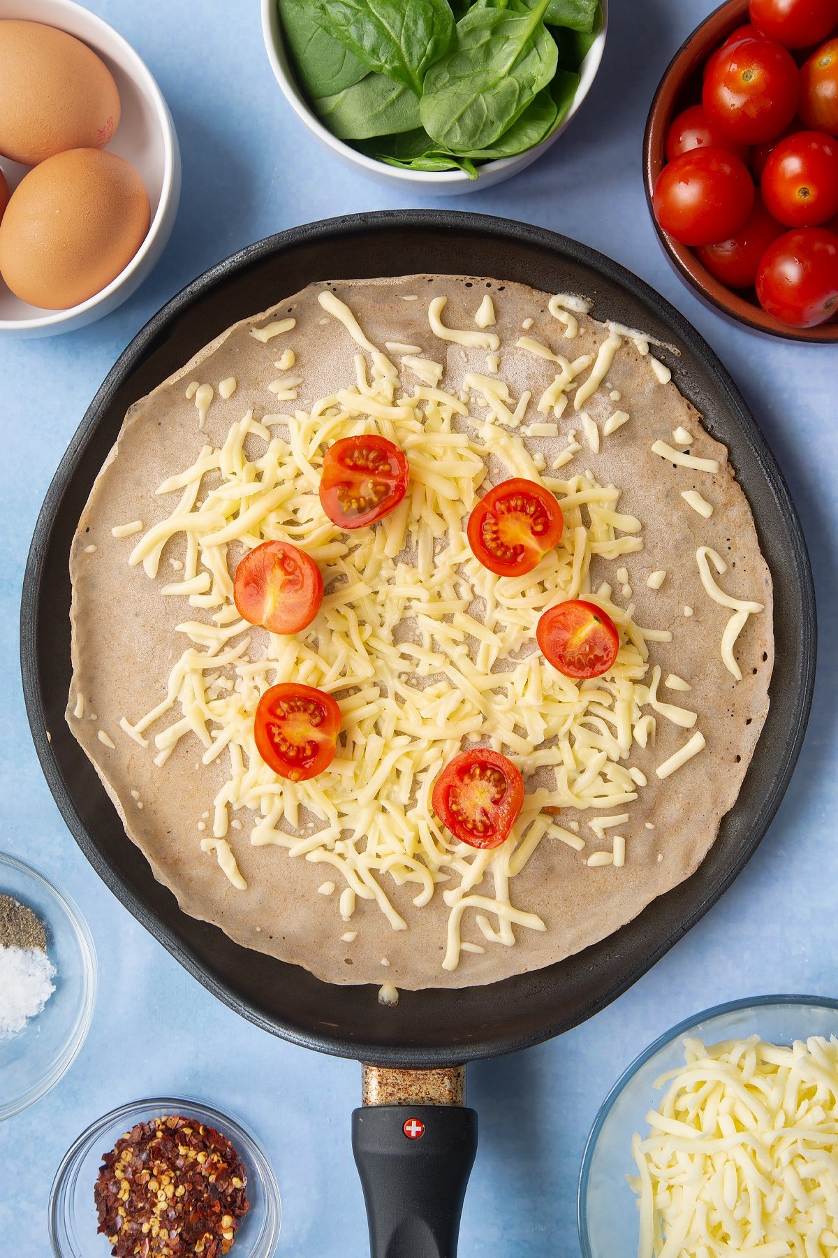 Buckwheat crepe topped with grated cheese and cherry tomatoes in a crepe pan. Ingredients to make buckwheat galettes surround the pan.